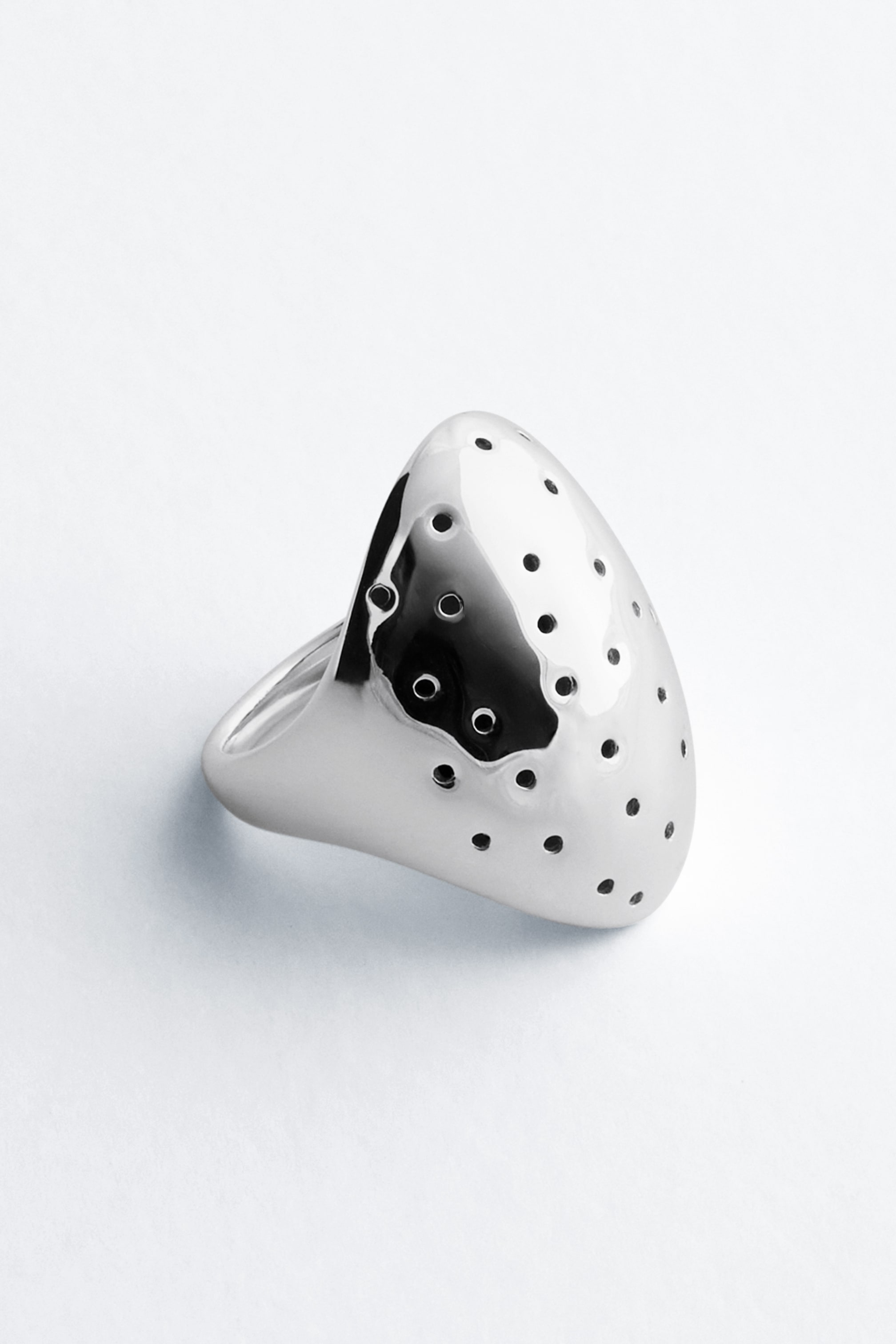 Agrandir l'image: La bague en laiton ajourée, dotée d’une finition polie ton argent, repose selon un angle de trois quarts. La bague met en valeur un large plateau bombé, en forme de bouclier, orné de petites perforations circulaires ajourées et se rétrécissant en un anneau lisse et épais.