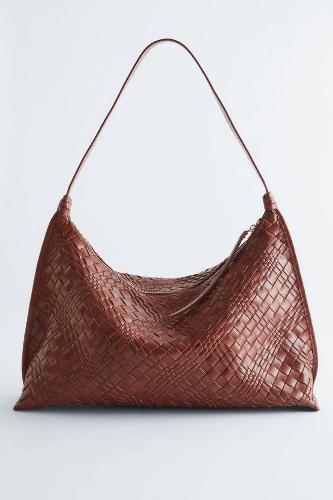 Een bruine leren tote bag wordt van de voorkant gepresenteerd, met een lichaam gemaakt van in elkaar gevlochten leren banden voor een gevlochten uitstraling. Het is ontworpen met een gebogen bovenkant en een ritssluiting, die is afgewerkt met een lange leren trekker. - 1