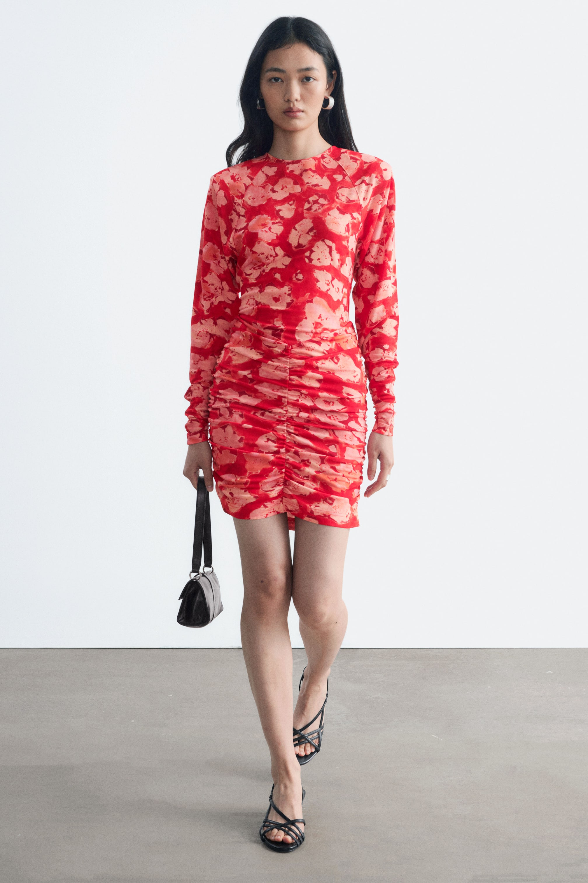 Vezi imaginea mai mare: O rochie mini cu fronseuri, cu un model floral abstract în nuanțe de roșu și portocaliu, cu mâneci lungi și decolteu rotund, este purtată de model. Rochia are fronseuri pronunțate pe partea din față a fustei mulate și este accesorizată cu o geantă mică neagră ținută într-o mână și sandale negre cu barete.