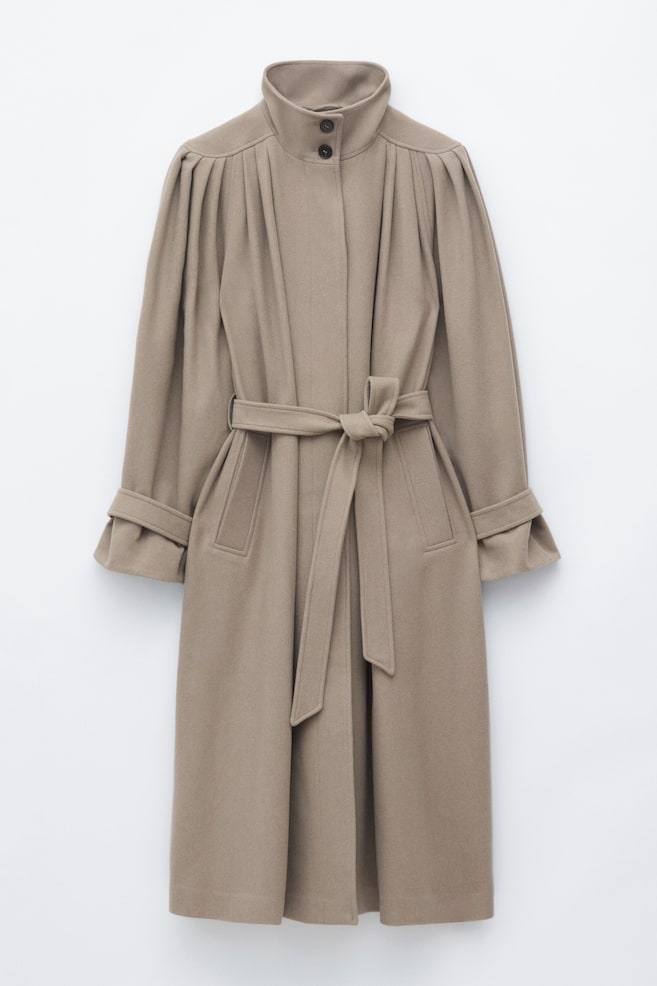 A taupe-coloured coat is shown front-facing, featuring a high funnel-neck collar with two dark buttons and prominent pleating details at the shoulders. The coat is styled with a matching self-tie belt knotted at the waist, diagonal welt pockets, and long sleeves accented with adjustable cuff straps. - 1