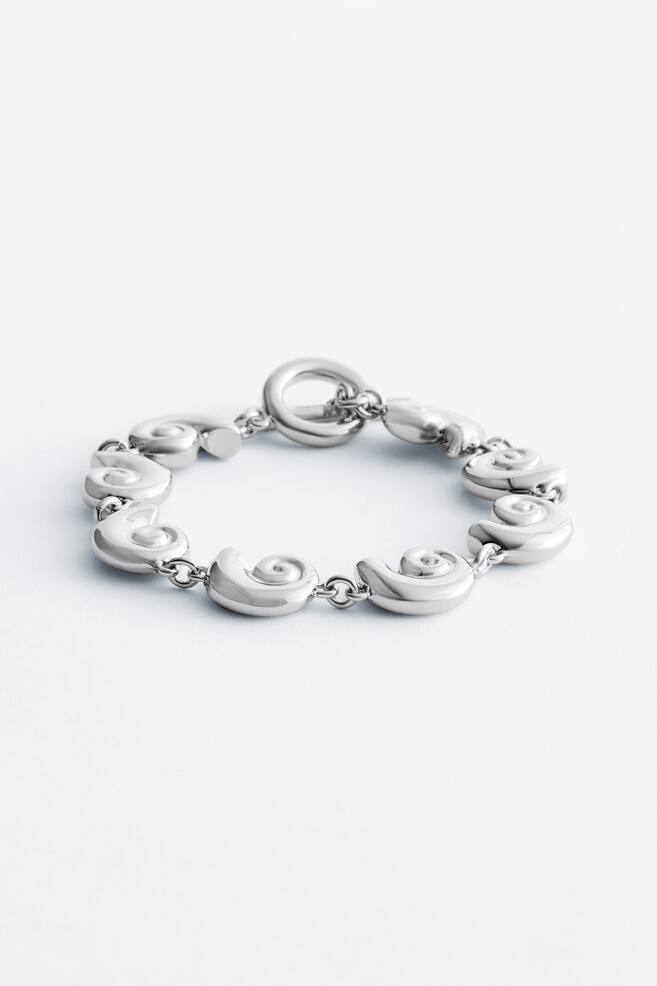 A silver-toned bracelet featuring multiple polished, spiral-shaped links, connected by small chain links, and fastening with a toggle clasp. It is lying flat, displaying its circular form from a front perspective. - 1