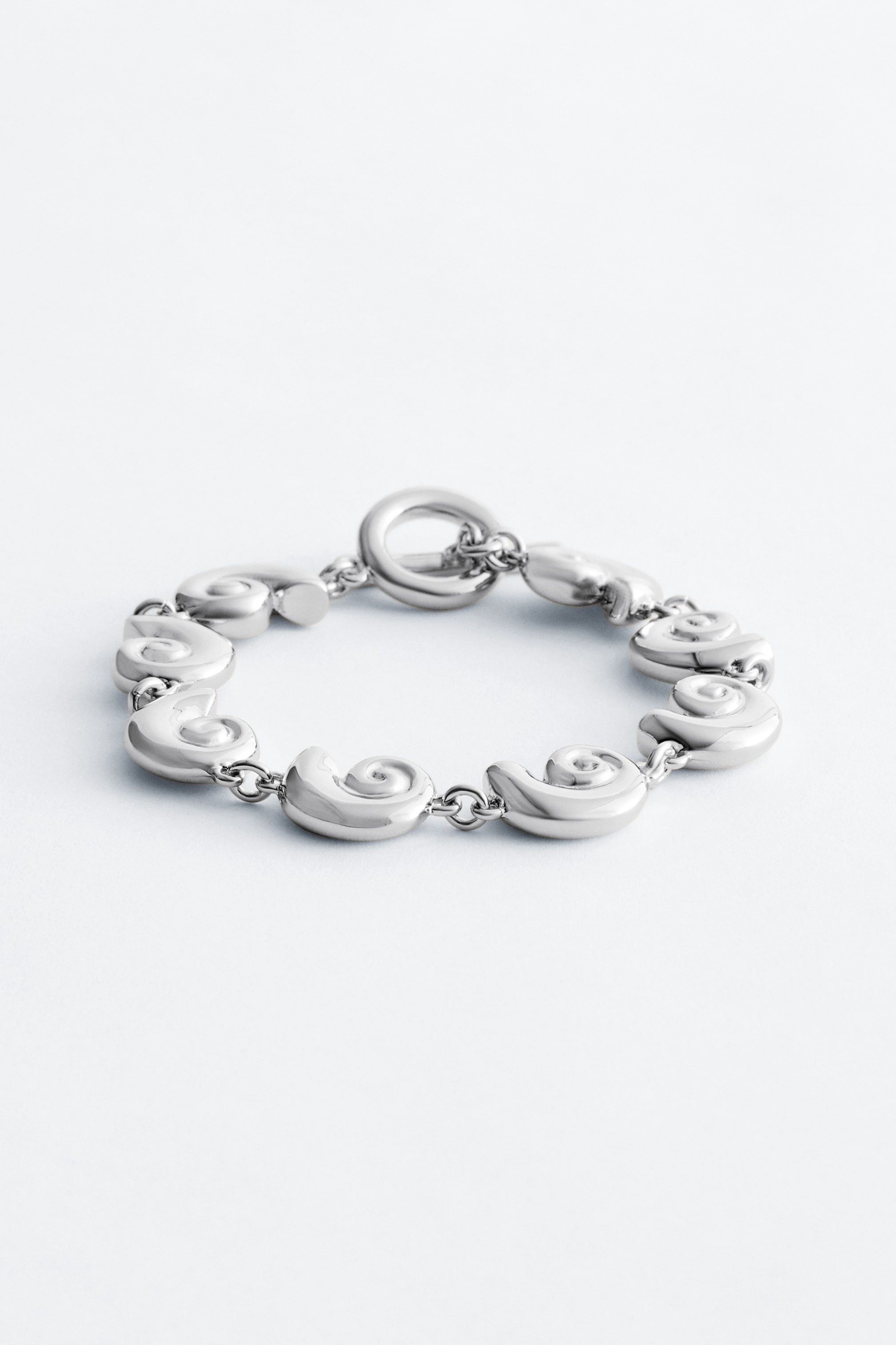 Grotere afbeelding bekijken: Een zilverkleurige armband met meerdere gepolijste, spiraalvormige schakels, verbonden door kleine kettingschakels en sluitend met een staafsluiting. Het ligt plat en toont zijn cirkelvorm vanuit een vooraanzicht.