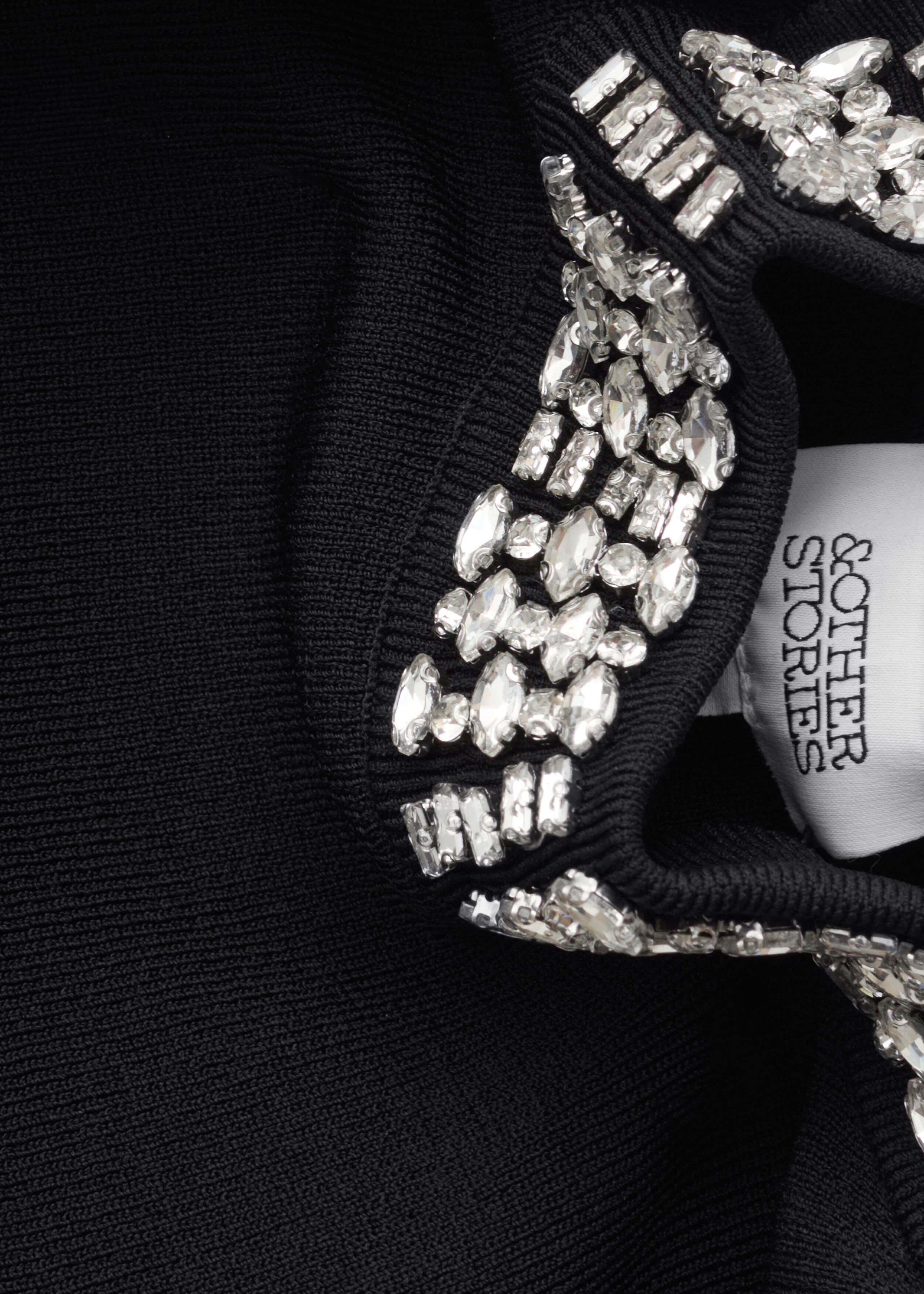 A black knit garment features a ribbed collar embellished with dense clusters of clear, faceted rhinestones in round, marquise, and rectangular shapes. A white woven brand label is visible on the inside of the neckline.