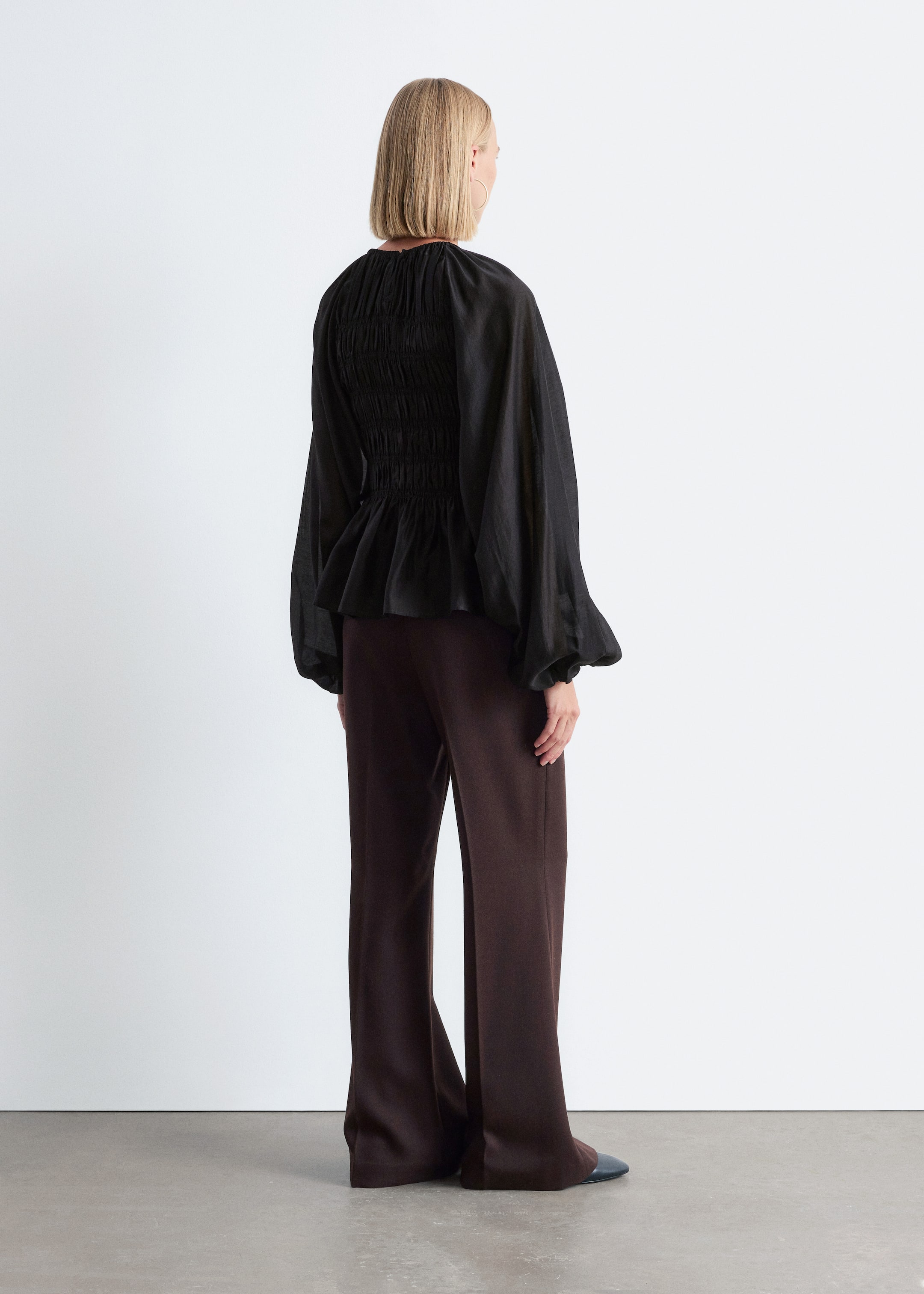 Grösseres Bild ansehen: Ein schwarzes Peplum-Top ist von hinten zu sehen, mit einem gesmokten Rückenteil und voluminösen langen Ärmeln mit elastischen Bündchen. Das Top fließt über dunkelbraune, weit geschnittene Hosen, die bis zum Boden reichen.