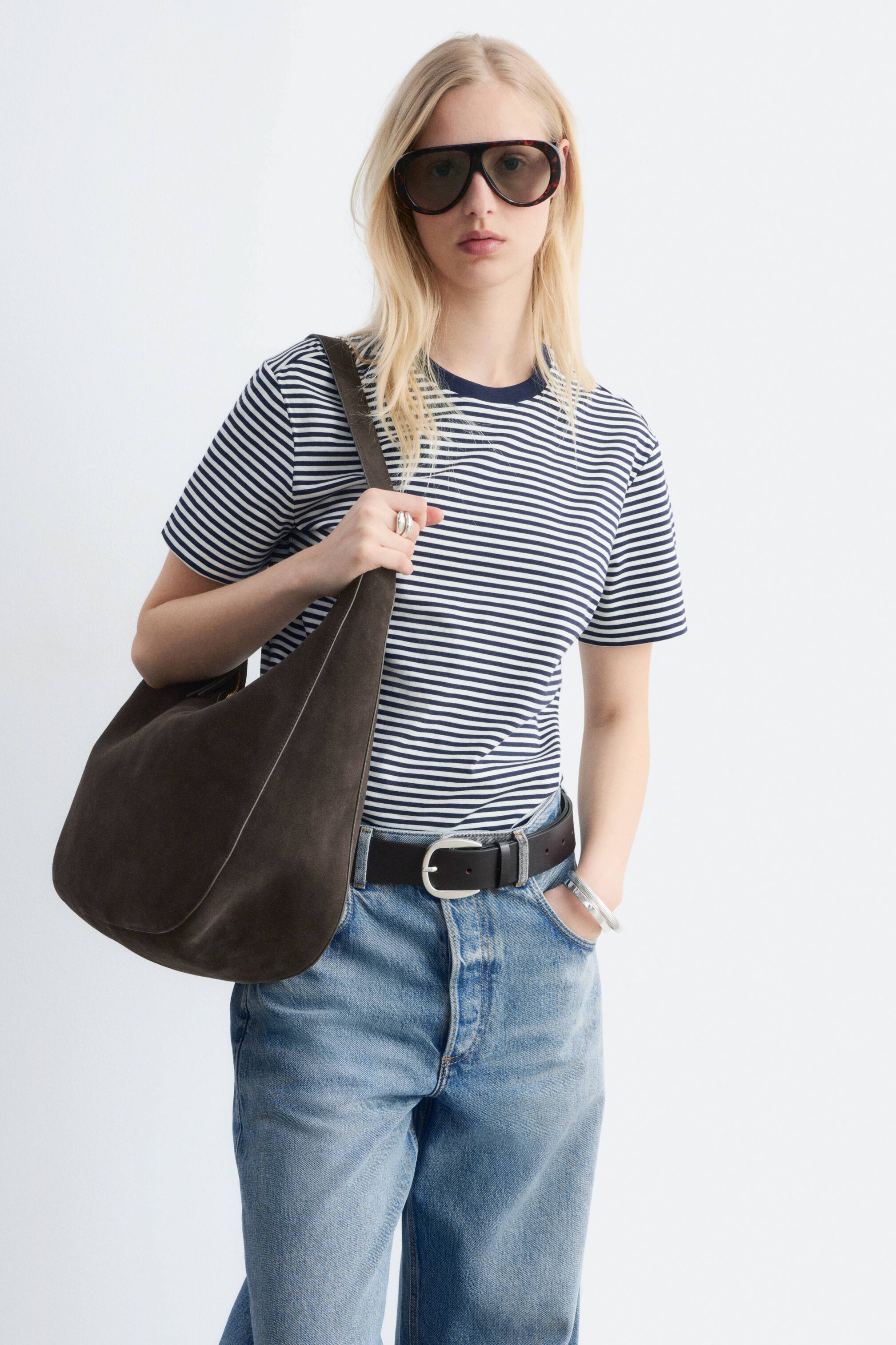 Größeres Bild anzeigen: Dunkelbraune Wildleder-Schultertasche hängt von der linken Schulter der Trägerin und zeigt sich von vorne. Sie präsentiert eine weiche, lässige Form mit einer hellen Nahtlinie, die entlang ihrer Kante sichtbar ist.