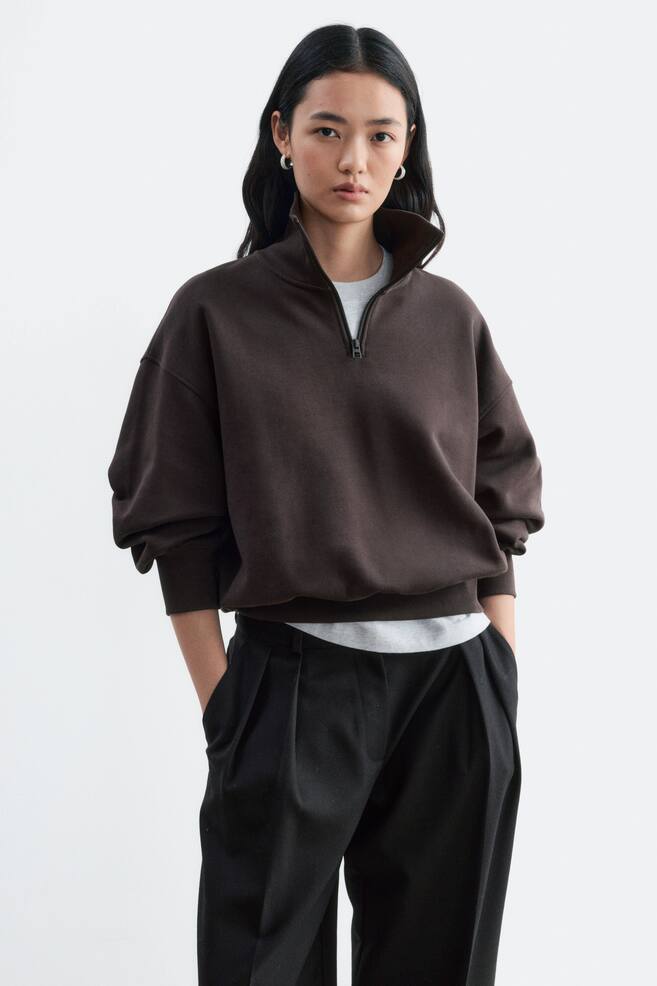 Ein dunkelbrauner Sweatshirt aus Baumwolljersey mit Half-Zip-Kragen und RippabschlÃ¼ssen wird vom Model getragen. Das Sweatshirt in Relaxed Fit wird Ã¼ber einem hellgrauen T-Shirt getragen und mit einer schwarzen Hose kombiniert. - 1