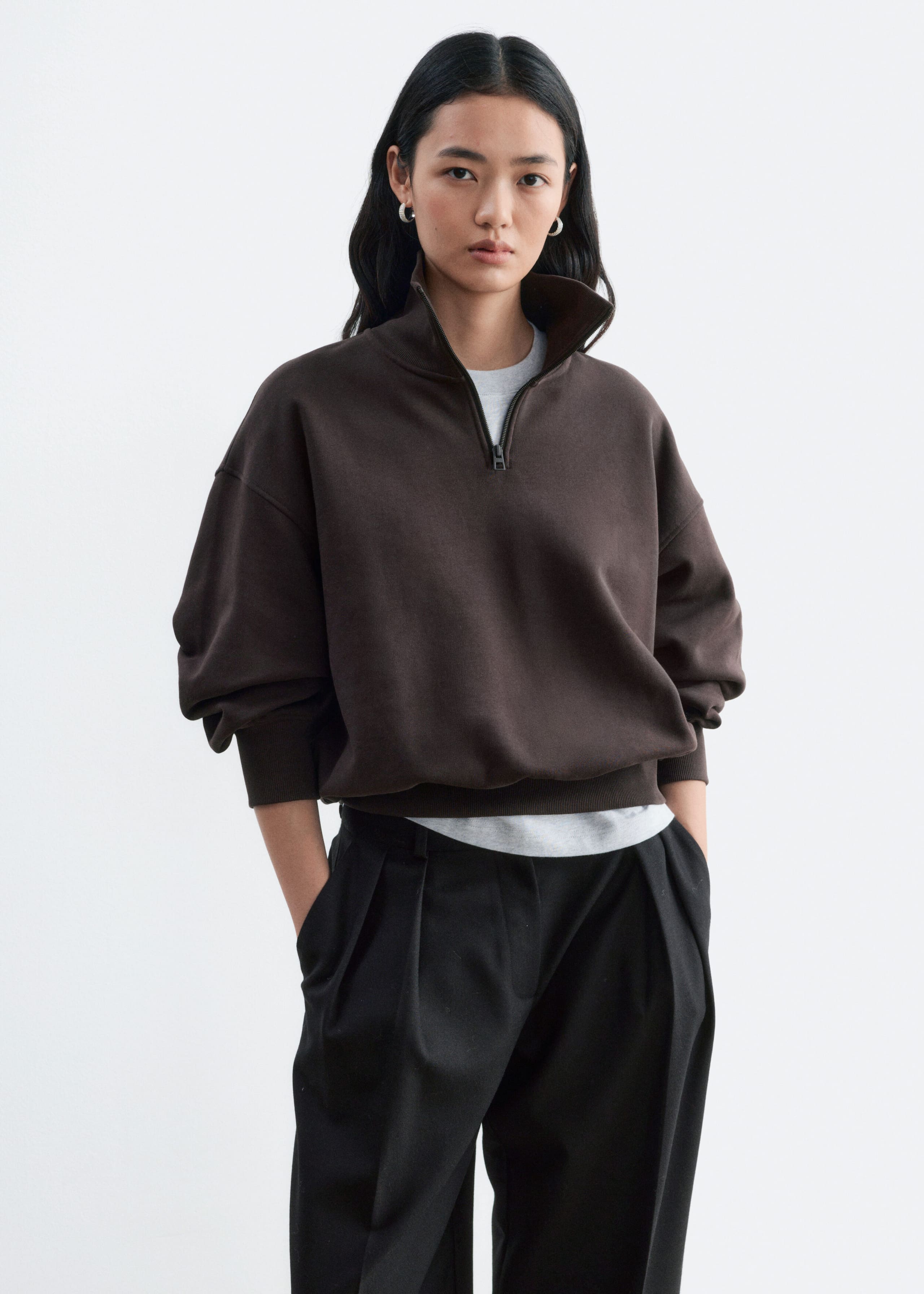 A dark brown cotton jersey sweatshirt featuring a half-zip collar and ribbed trims is being worn by the model. The relaxed-fit sweatshirt is layered over a light grey T-shirt and paired with black trousers.