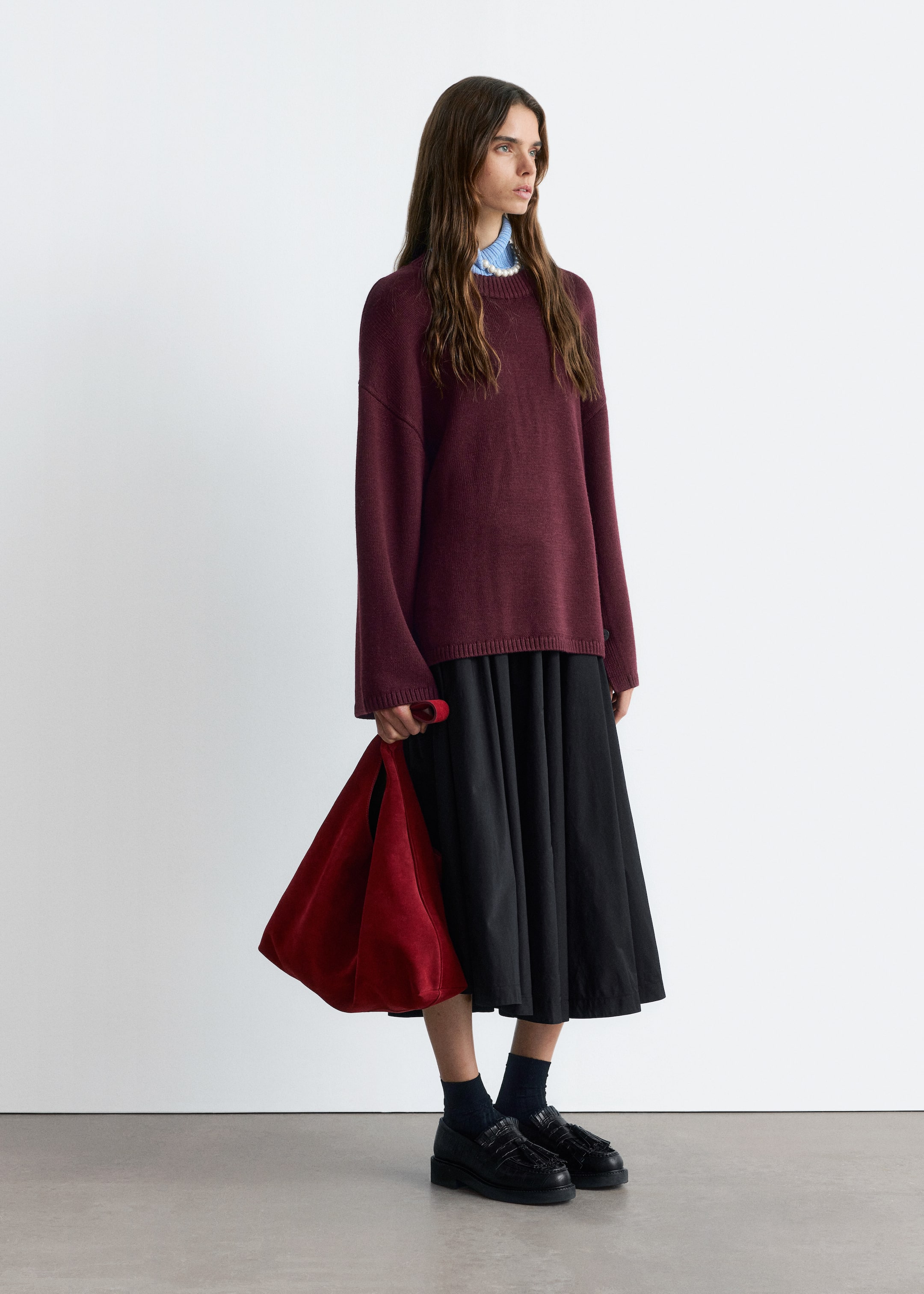 View larger image: A burgundy oversized crew-neck sweater is being displayed from a slight right angle, revealing its relaxed fit as it layers over a light blue collared shirt and a pearl necklace. It is being paired with a black pleated midi skirt, black chunky loafers featuring fringe and dark socks, and a red triangular suede bag is being held at the side.