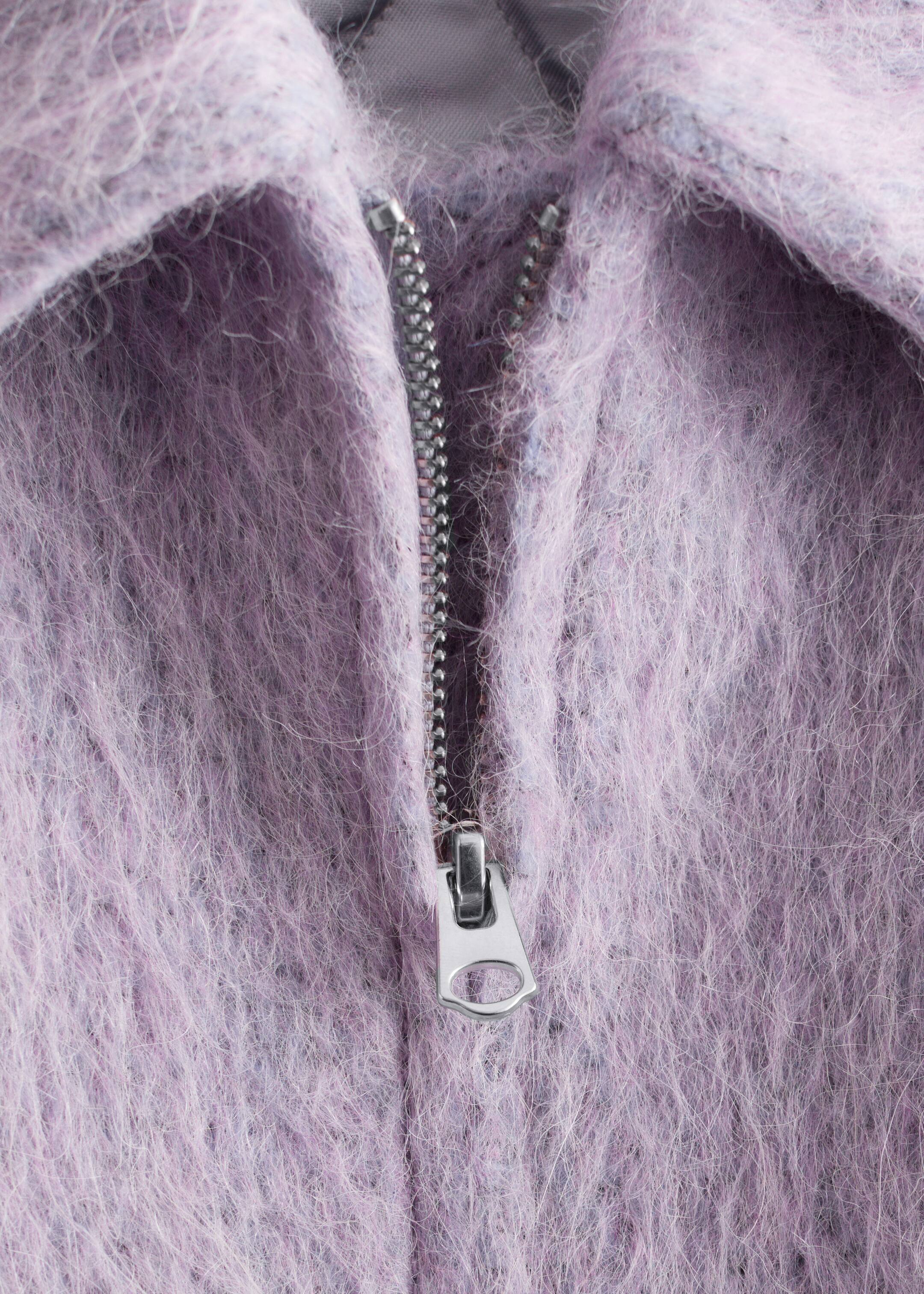 Visualizza immagine più grande: Un indumento di colore viola chiaro, caratterizzato da una texture prominente a pelo lungo, presenta una chiusura a zip di tonalità argento parzialmente aperta lungo la parte anteriore, con un colletto piegato visibile nella parte superiore.