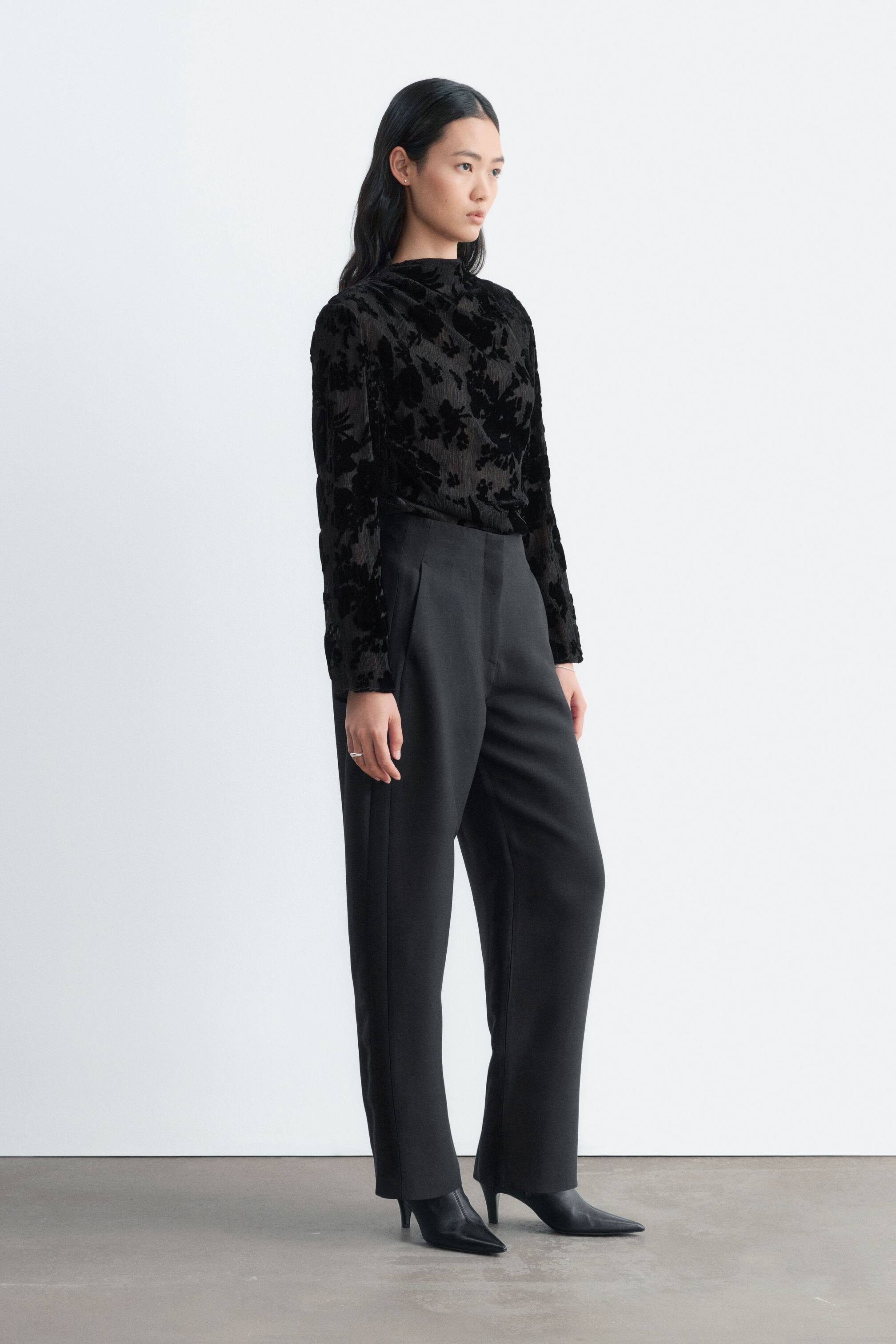 View larger image: A black long-sleeved blouse with a textured velvet floral pattern is tucked into high-waisted black barrel-leg trousers. The model is standing in a three-quarter profile, showcasing the relaxed drape of the top and the structured silhouette of the trousers.