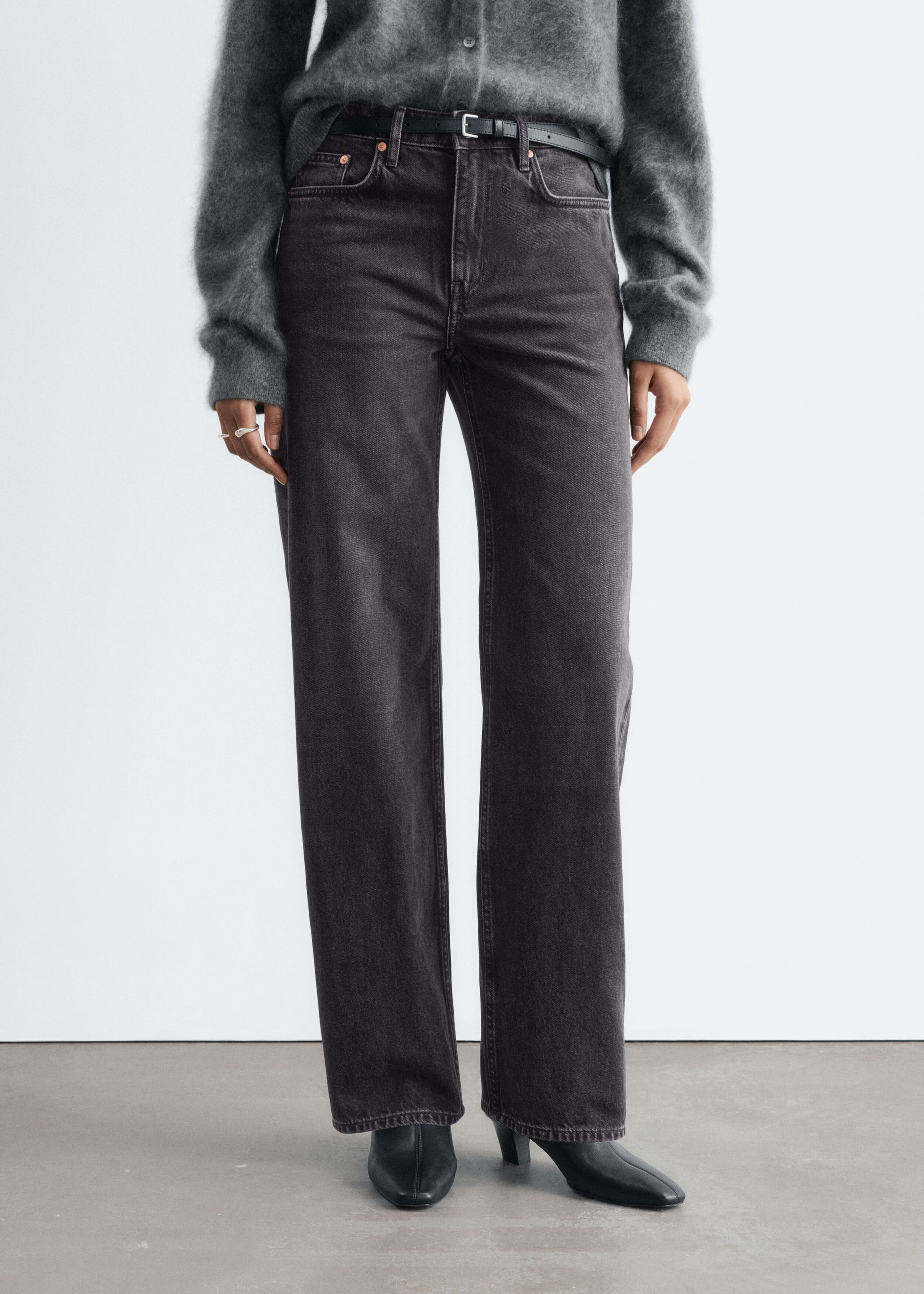 The model is standing and wearing straight-leg, mid-rise jeans in washed dark gray denim. The jeans feature a classic five-pocket design and are styled with a thin black belt, a gray knit cardigan, and black pointed-toe boots.