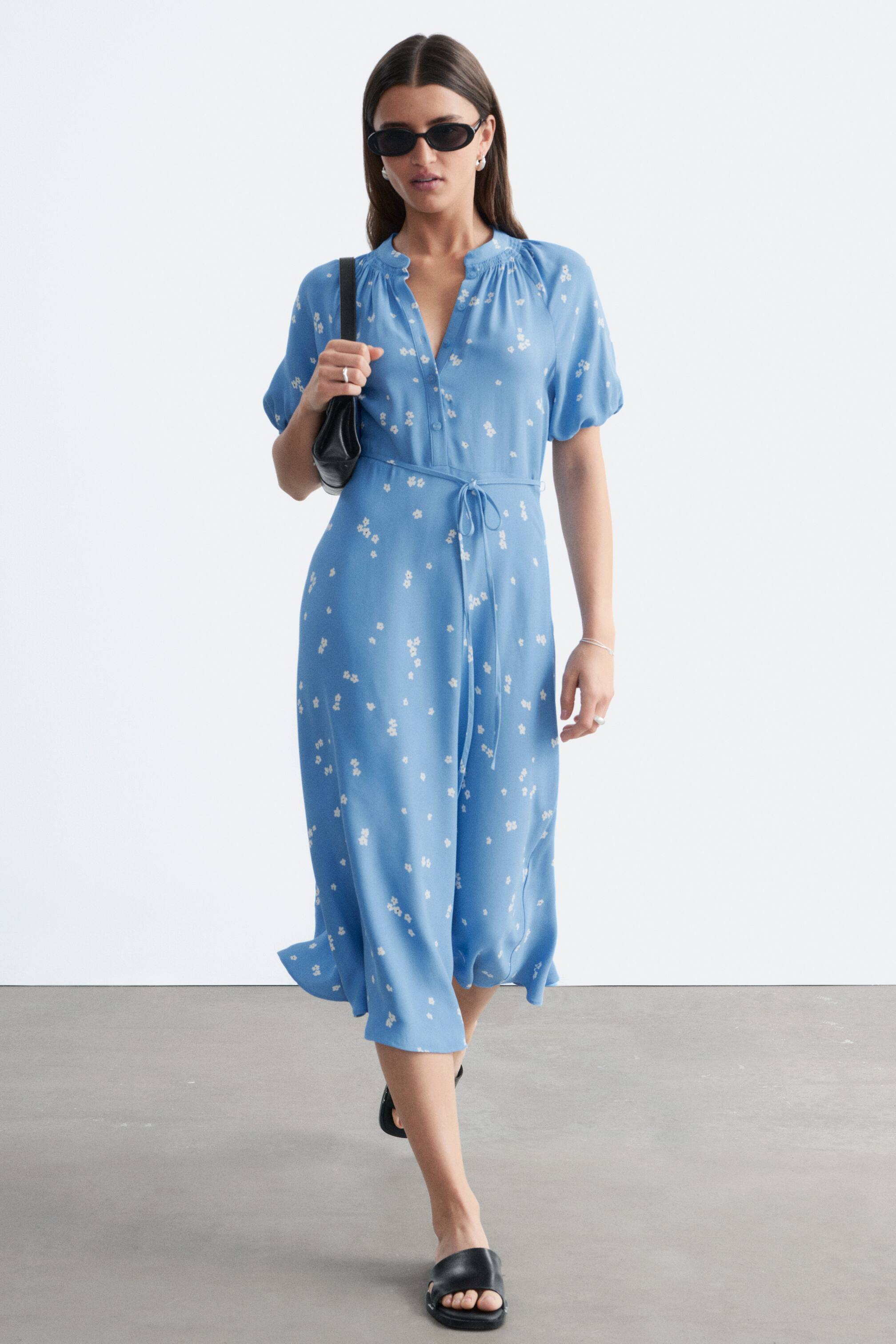 Agrandir l'image: Une robe midi bleu clair ornée de petits motifs floraux blancs, dotée d'un col en V avec un col froncé, de manches courtes bouffantes et d'une ceinture à nouer à la taille, mettant en valeur sa silhouette fluide vue sous un léger angle de face.
