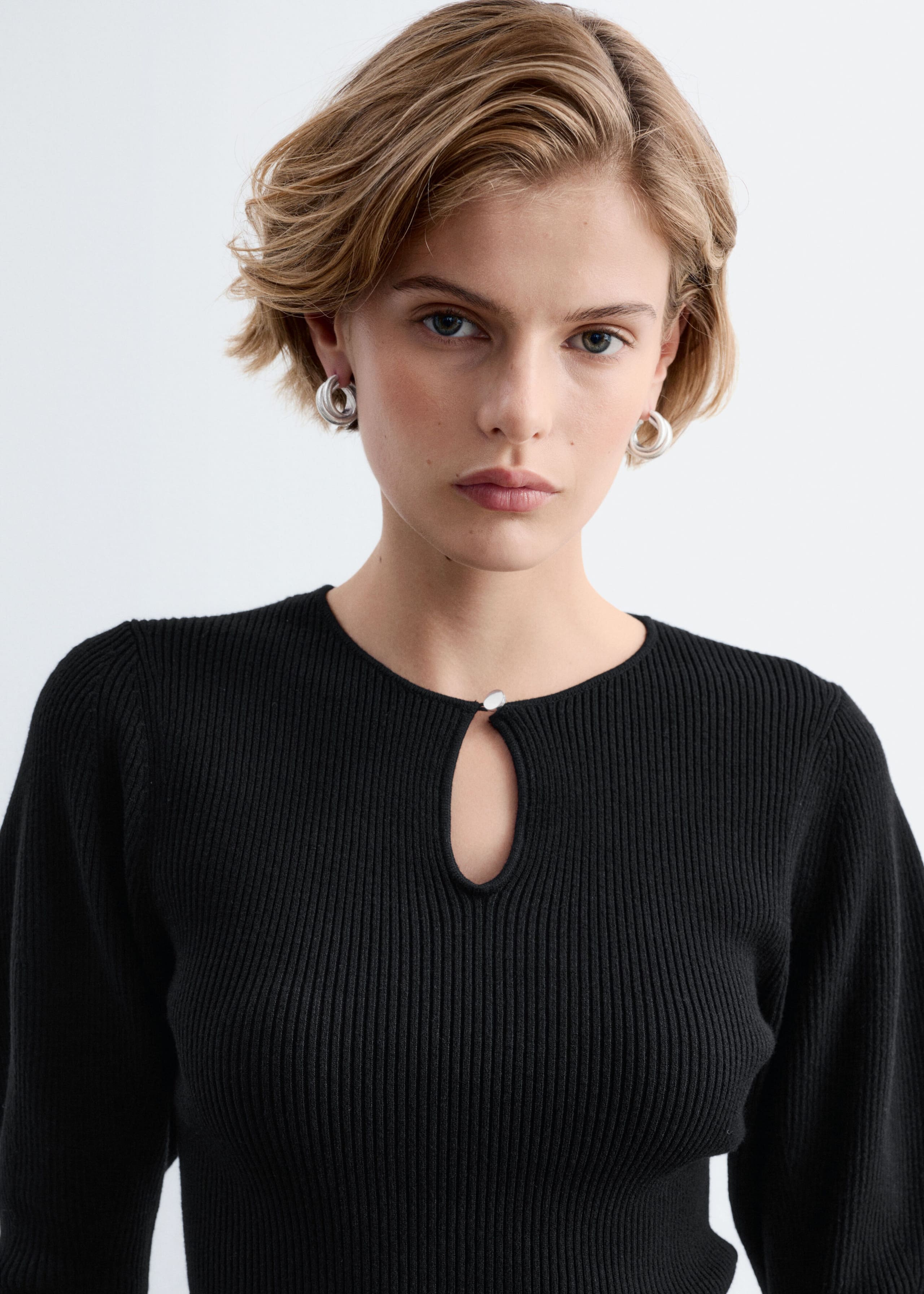 The model is wearing a black rib-knit top featuring a keyhole opening at the front neckline secured with a small silver button. The slim-fitting top has a crew neckline and slightly puffed shoulders.
