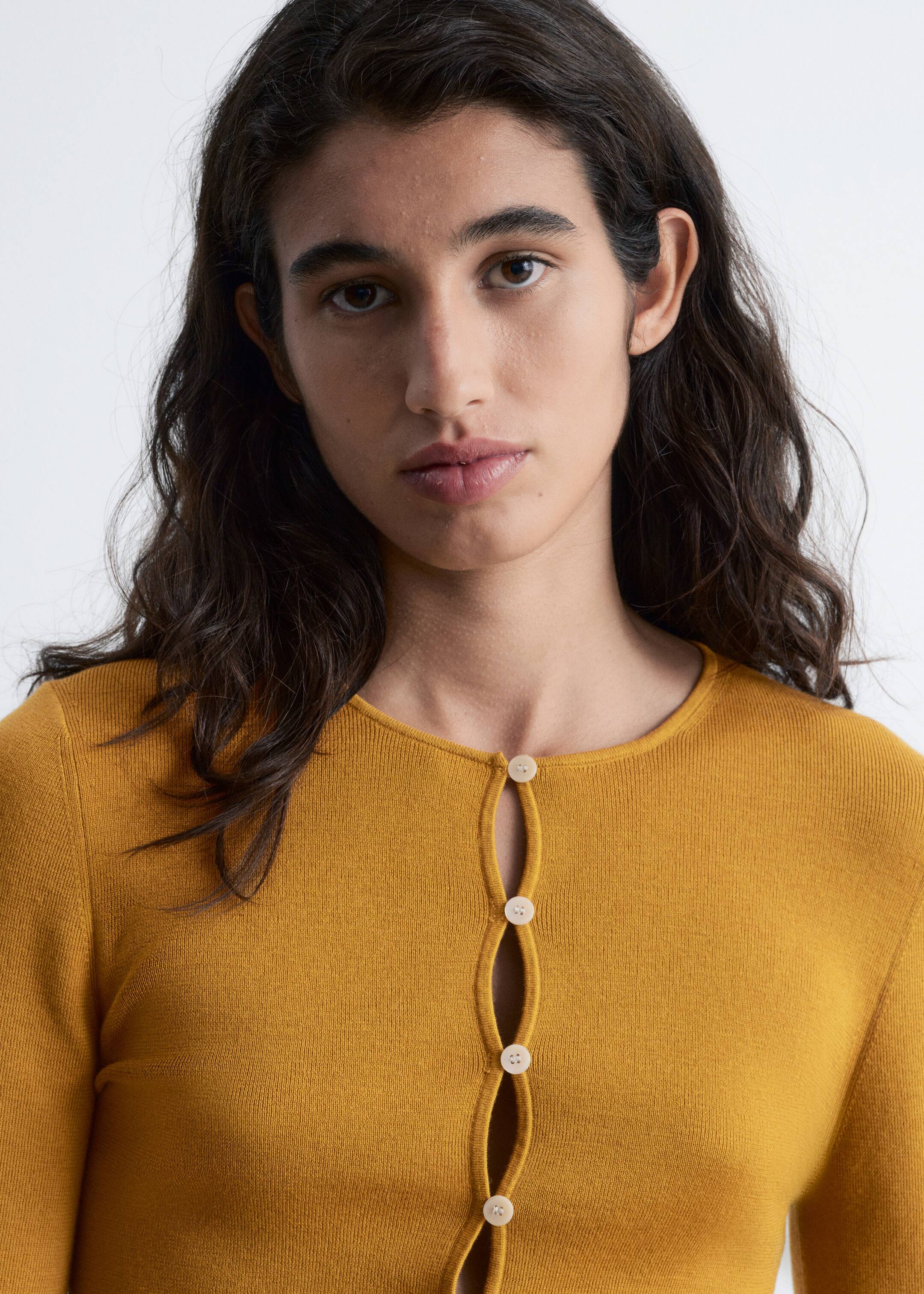 Agrandir l'image: Un haut en tricot jaune moutarde est porté, avec un col rond et des manches longues. Sa patte de boutonnage avant unique présente des boutons ronds de couleur claire, espacés pour créer un design ouvert ondulant au centre.