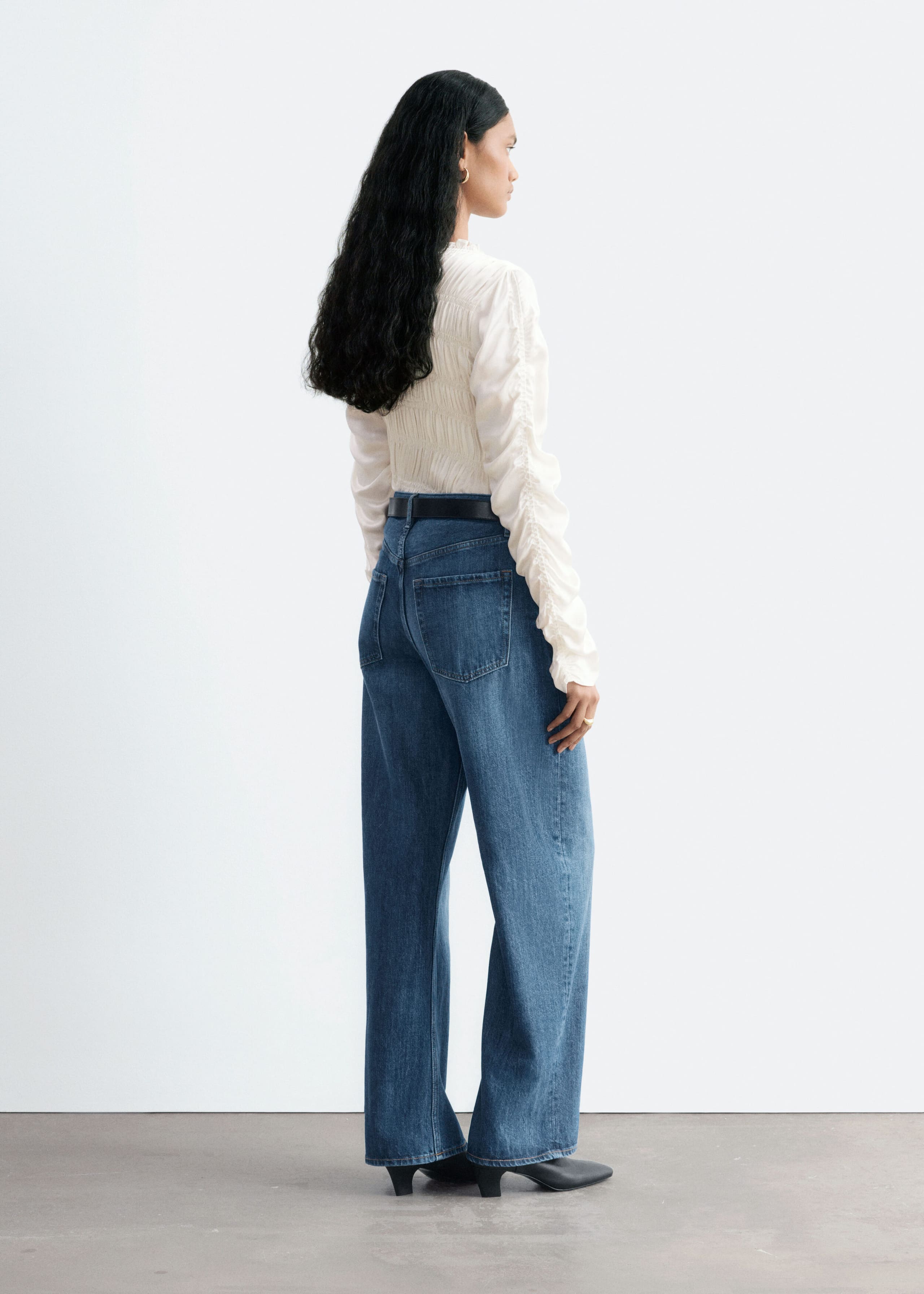 Blue wide-leg denim jeans are being worn by the model, styled with a black leather belt, a tucked-in textured white blouse, and black heeled shoes. The jeans feature a relaxed, oversized fit with visible back pockets and are shown from a three-quarter rear view.