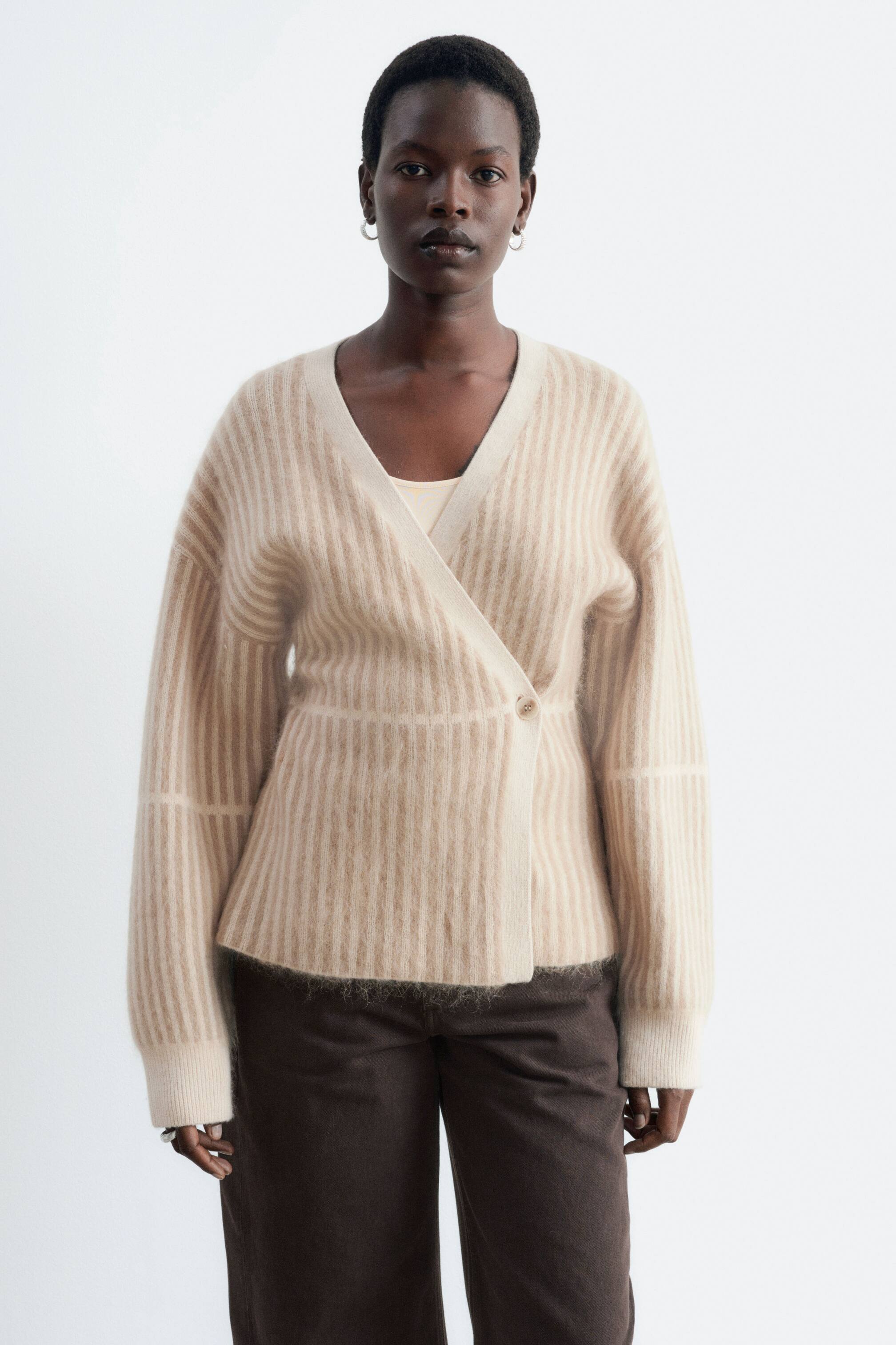 Vezi imaginea mai mare: Un cardigan petrecut, cu dungi în bej deschis și alb murdar, este purtat de model. Acest cardigan tricotat pufos prezintă un decolteu în V, închidere cu un singur nasture și modele cu dungi verticale și orizontale, iar dedesubt este purtat un top deschis la culoare, asortat cu pantaloni închiși la culoare.