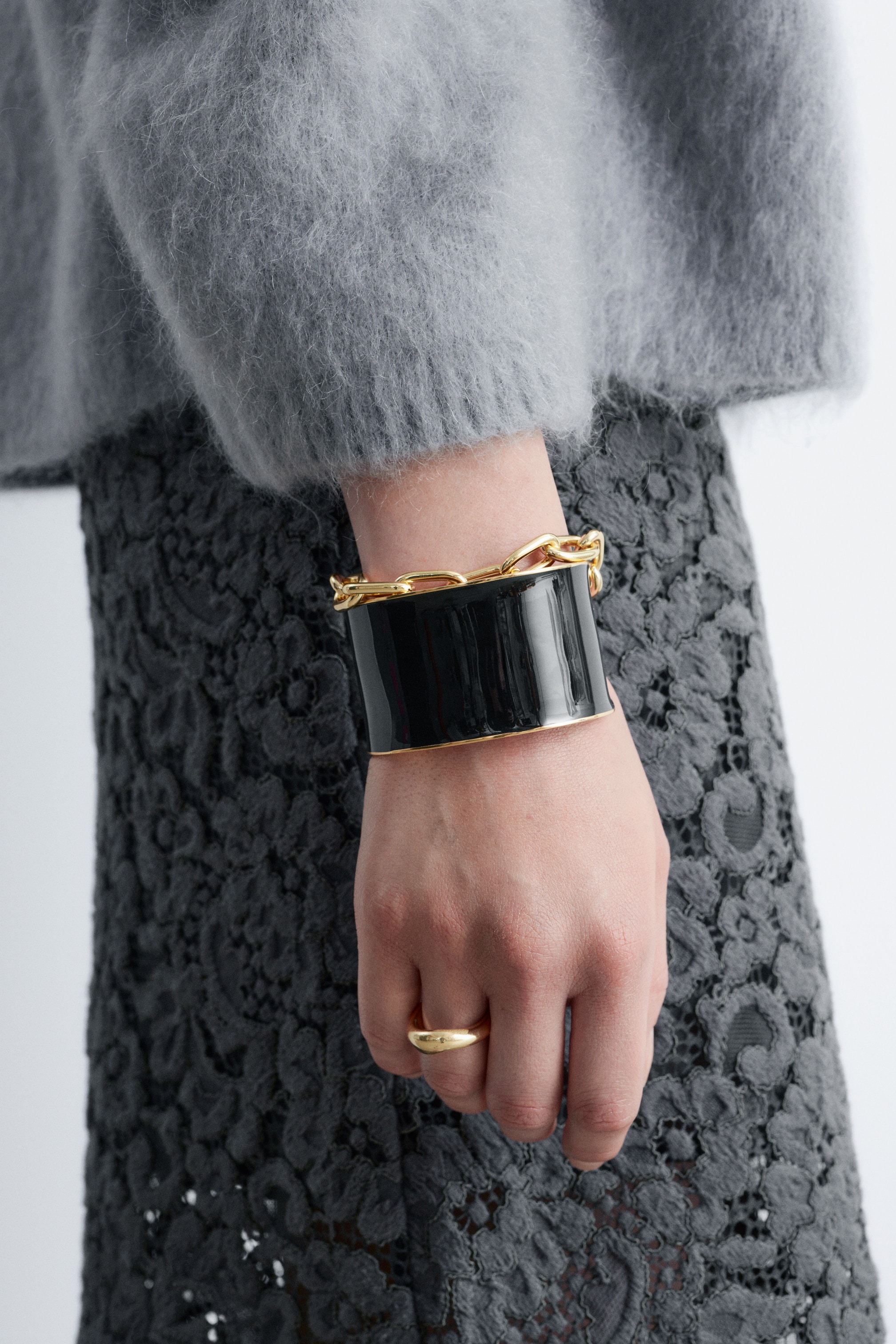 Agrandir l'image: Un large bracelet manchette noir, doté d'un bord intérieur doré, est porté au poignet du mannequin, avec un bracelet à maillons dorés superposé au-dessus.