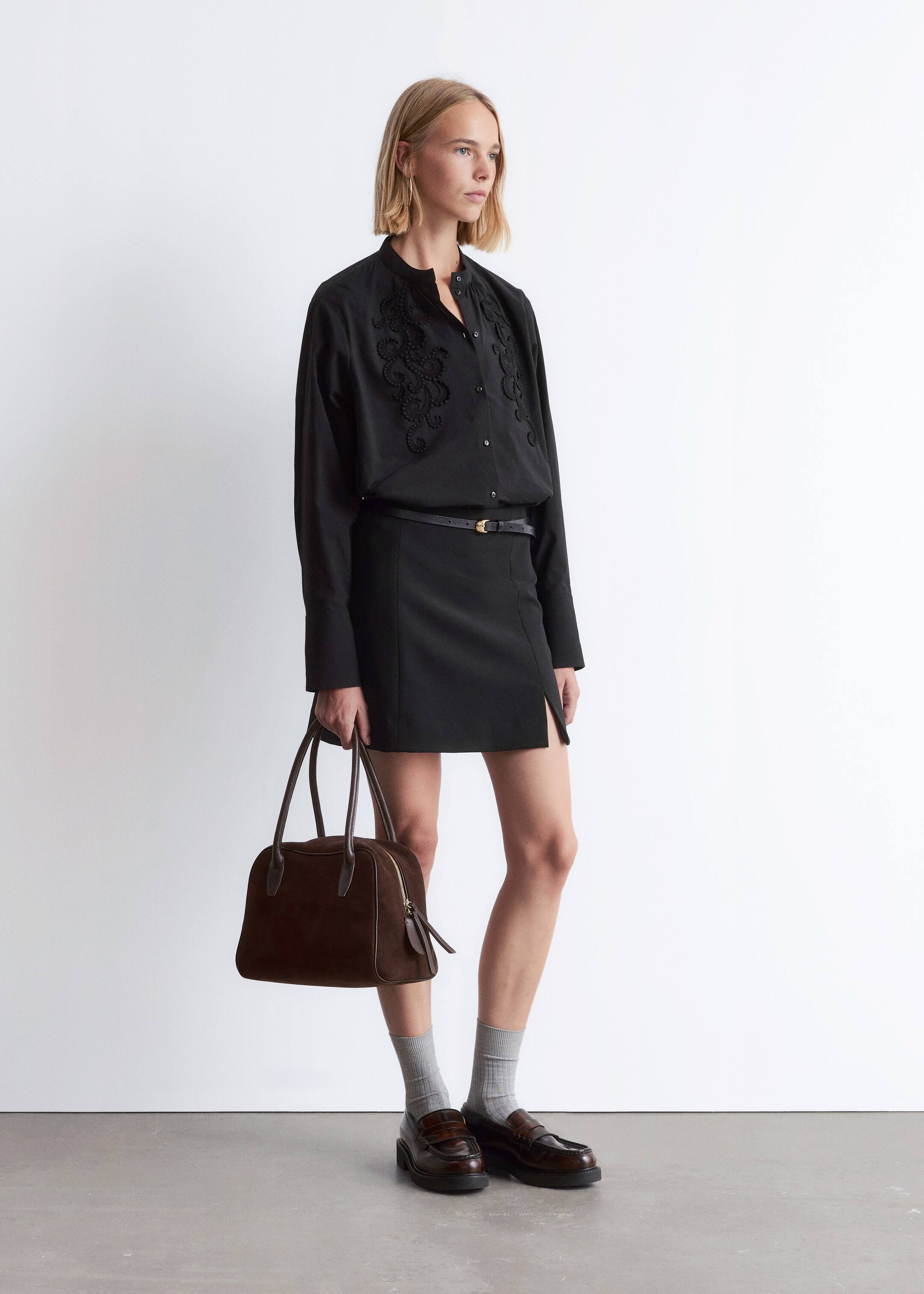 View larger image: A black long-sleeved shirt with intricate black embroidery on the upper chest and a band collar is being worn, tucked into a black mini skirt featuring a subtle front slit and cinched by a thin black belt with a gold-toned buckle. A dark brown structured handbag with two top handles and a visible zipper closure is being held, while light grey ribbed socks are peeking out above dark brown chunky penny loafers.