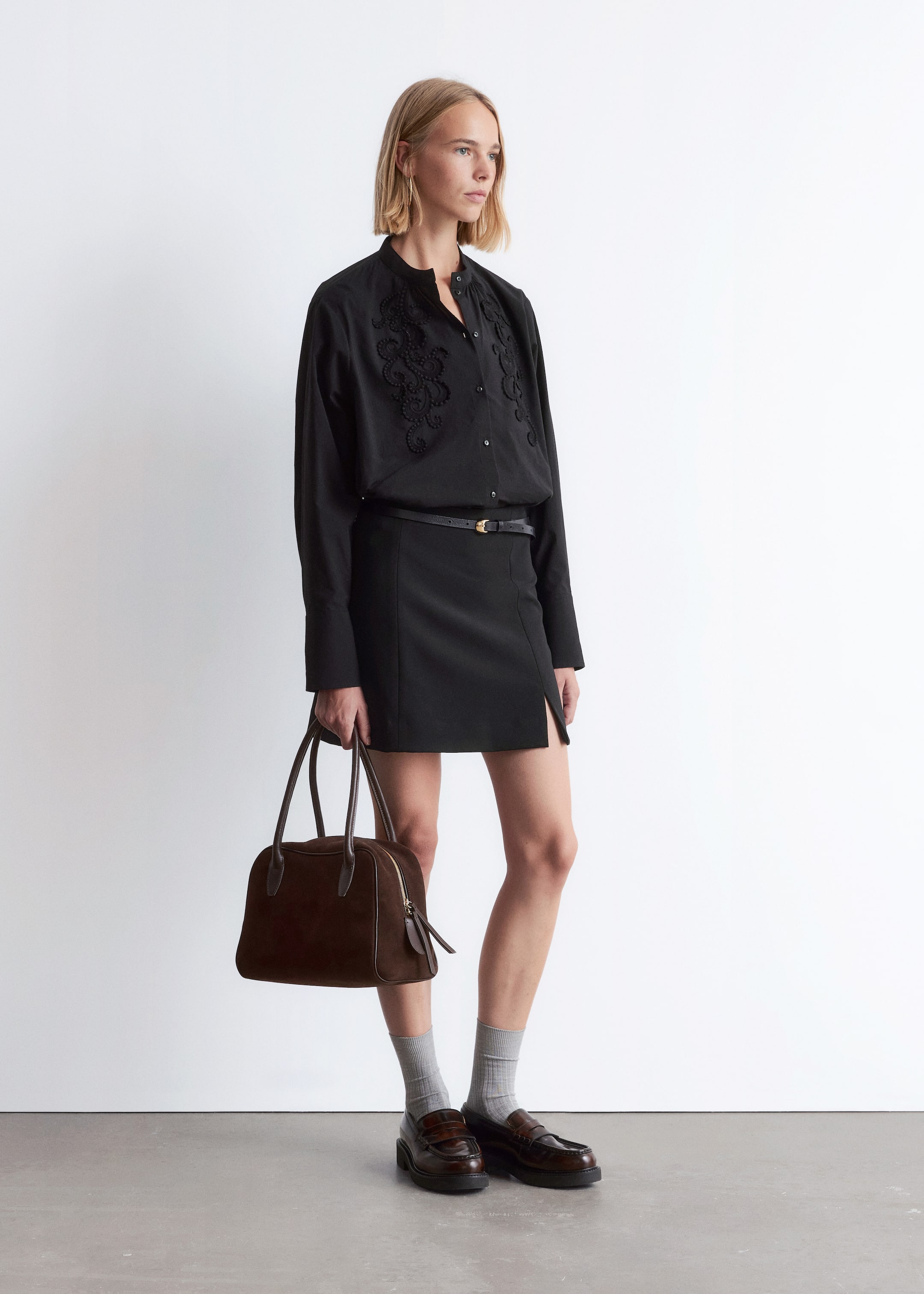 Agrandir l'image: Une chemise noire à manches longues avec une broderie noire complexe sur le haut de la poitrine et un col officier est portée, rentrée dans une mini-jupe noire avec une fente subtile à l'avant et cintrée par une fine ceinture noire avec une boucle dorée. Un sac à main structuré marron foncé avec deux poignées supérieures et une fermeture éclair visible est tenu, tandis que des chaussettes côtelées gris clair dépassent au-dessus de mocassins marron foncé à semelles épaisses.