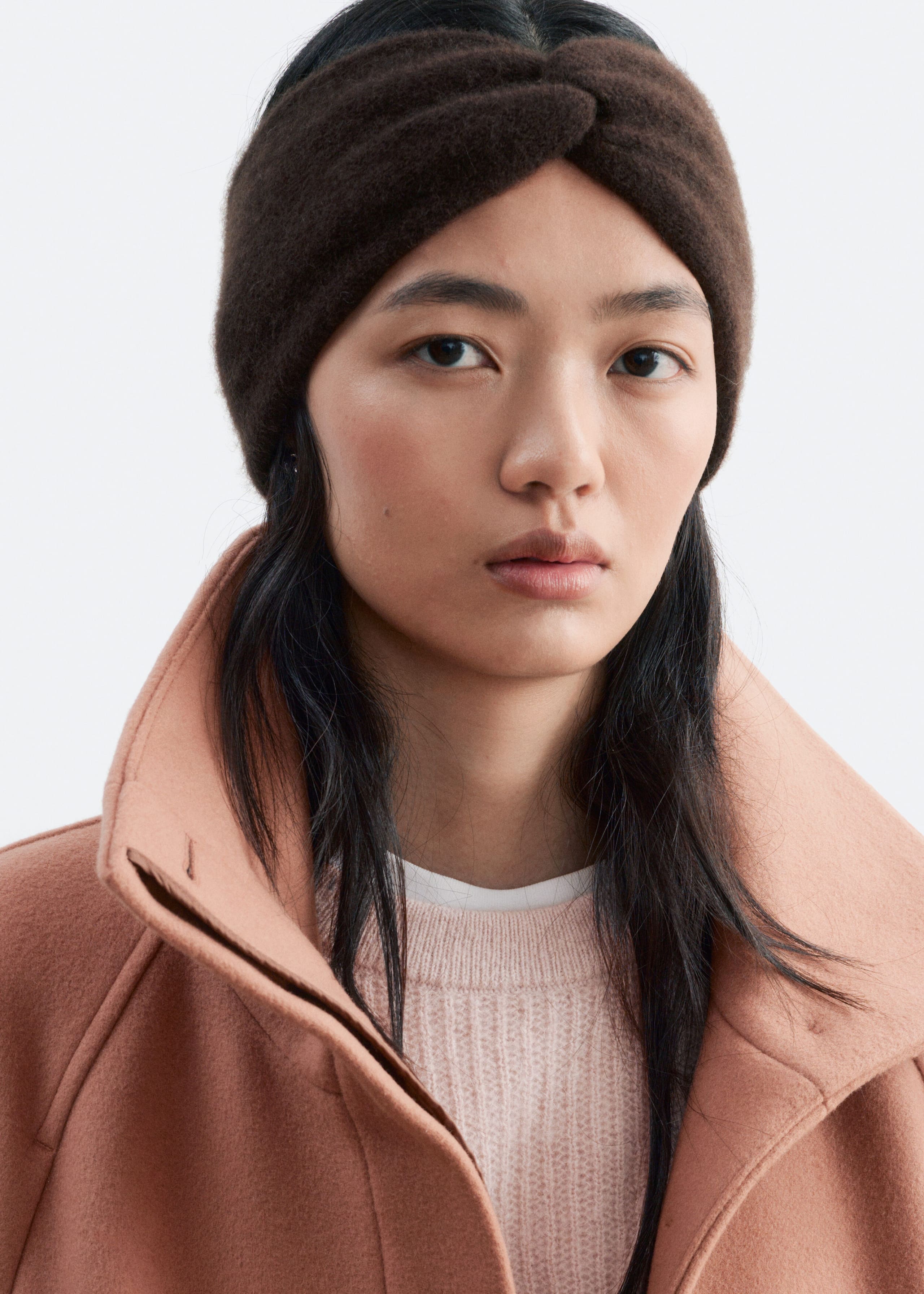A wide, dark brown knitted cashmere headband with a central twist knot is being worn on the model's head. The headband is styled with a light pink crew neck sweater and a terracotta-coloured coat.