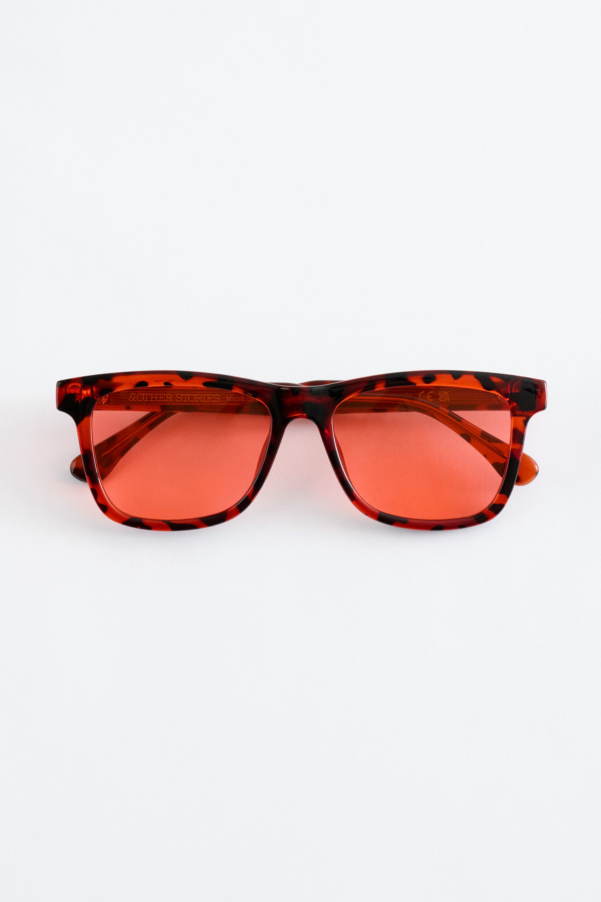 Ver imagem maior: Um par de óculos de sol está voltado para a frente, com uma armação de acetato em padrão tartaruga. É desenhado com um perfil retangular e bordas suavemente arredondadas, e as lentes têm um tom translúcido de vermelho-alaranjado.