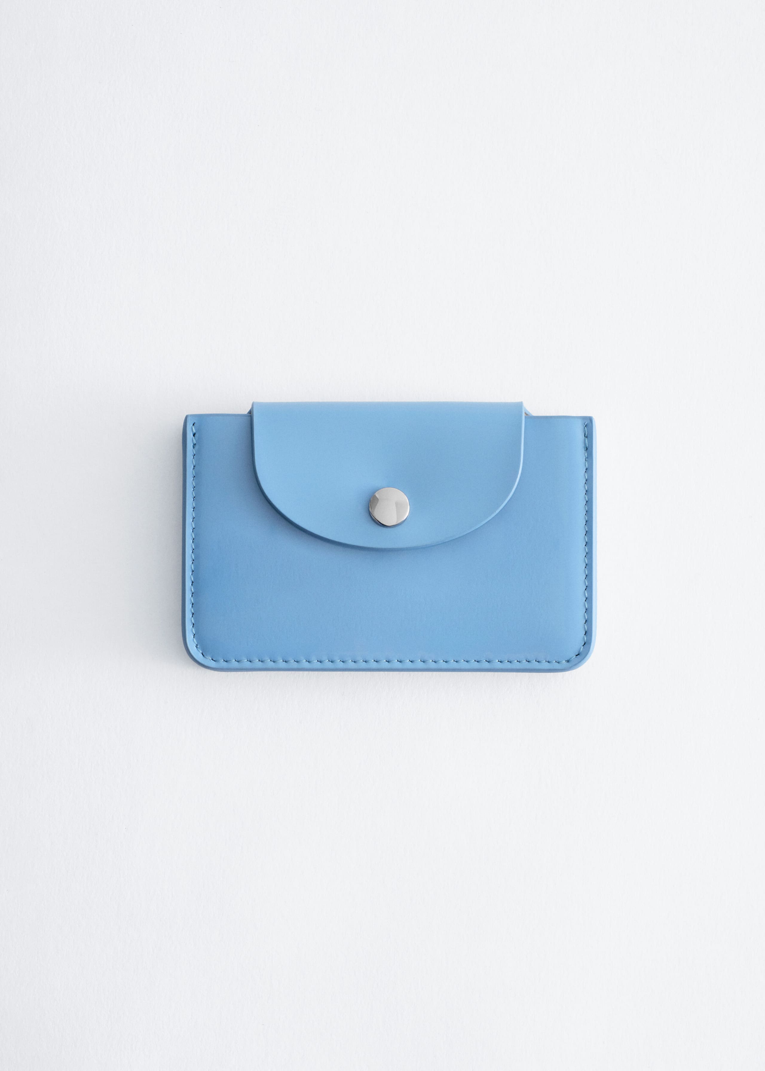 A light blue leather cardholder is shown facing forward, featuring a curved front flap with a silver-tone snap closure and tonal stitching along the edges.