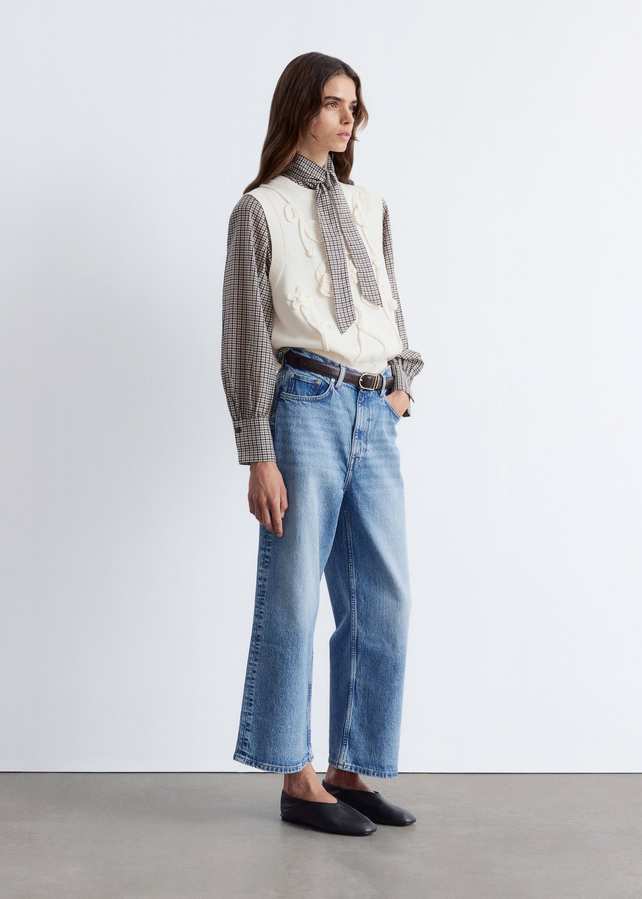 Größeres Bild ansehen: Ein Outfit besteht aus einer cremefarbenen Strickweste mit erhabenen Blumenmustern, die über einem braun-weiß karierten Langarmhemd mit hohem, gebundenem Kragen getragen wird. Das Hemd wird mit hell gewaschenen, weit geschnittenen Jeans kombiniert, die mit einem dunkelbraunen Gürtel geschnürt und mit schwarzen Slippern vervollständigt werden.