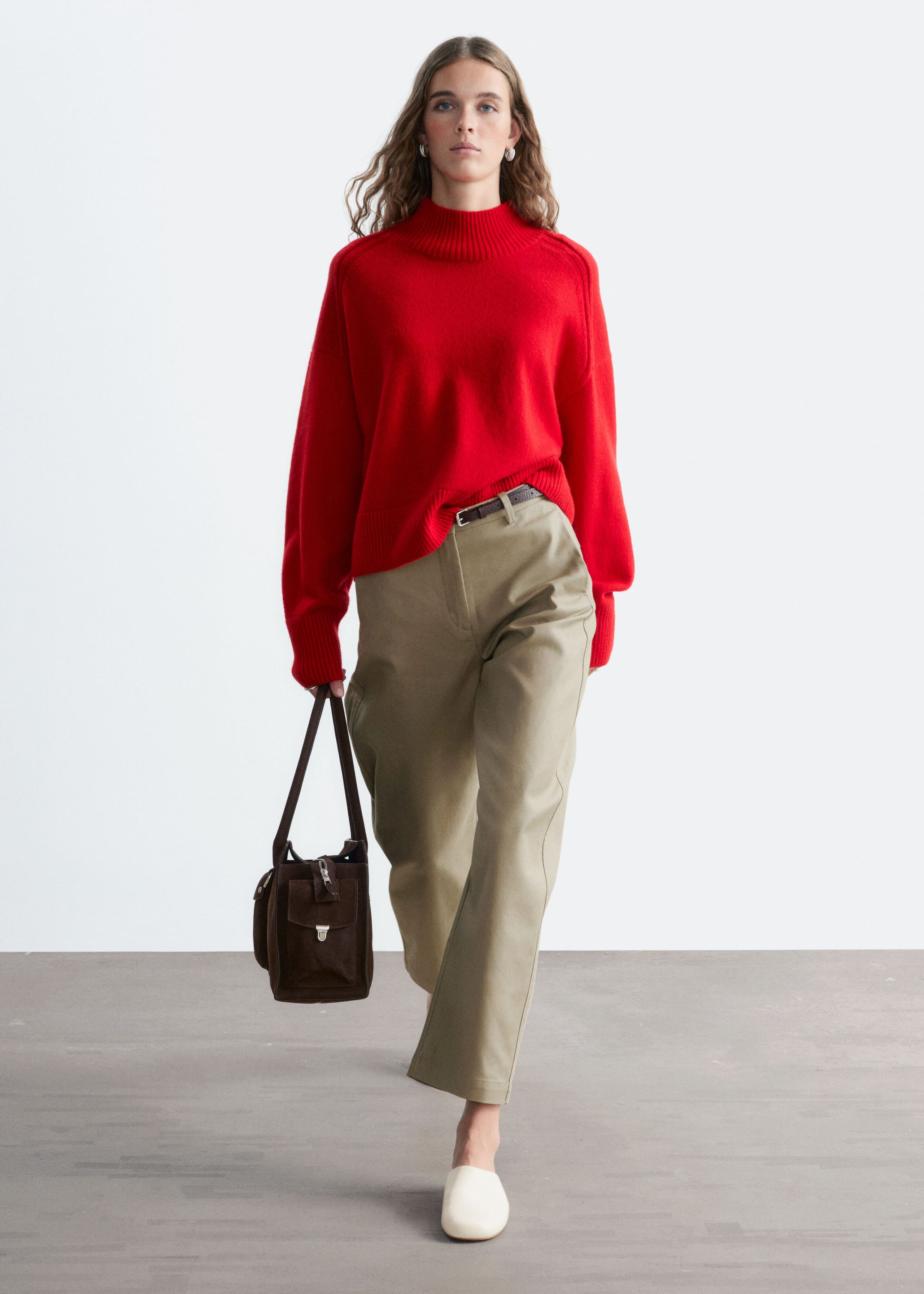 A model is wearing a red mock-neck sweater, with the front hem tucked into high-waisted khaki trousers. The long-sleeve knitwear features a relaxed fit and ribbed detailing on the high collar and cuffs.