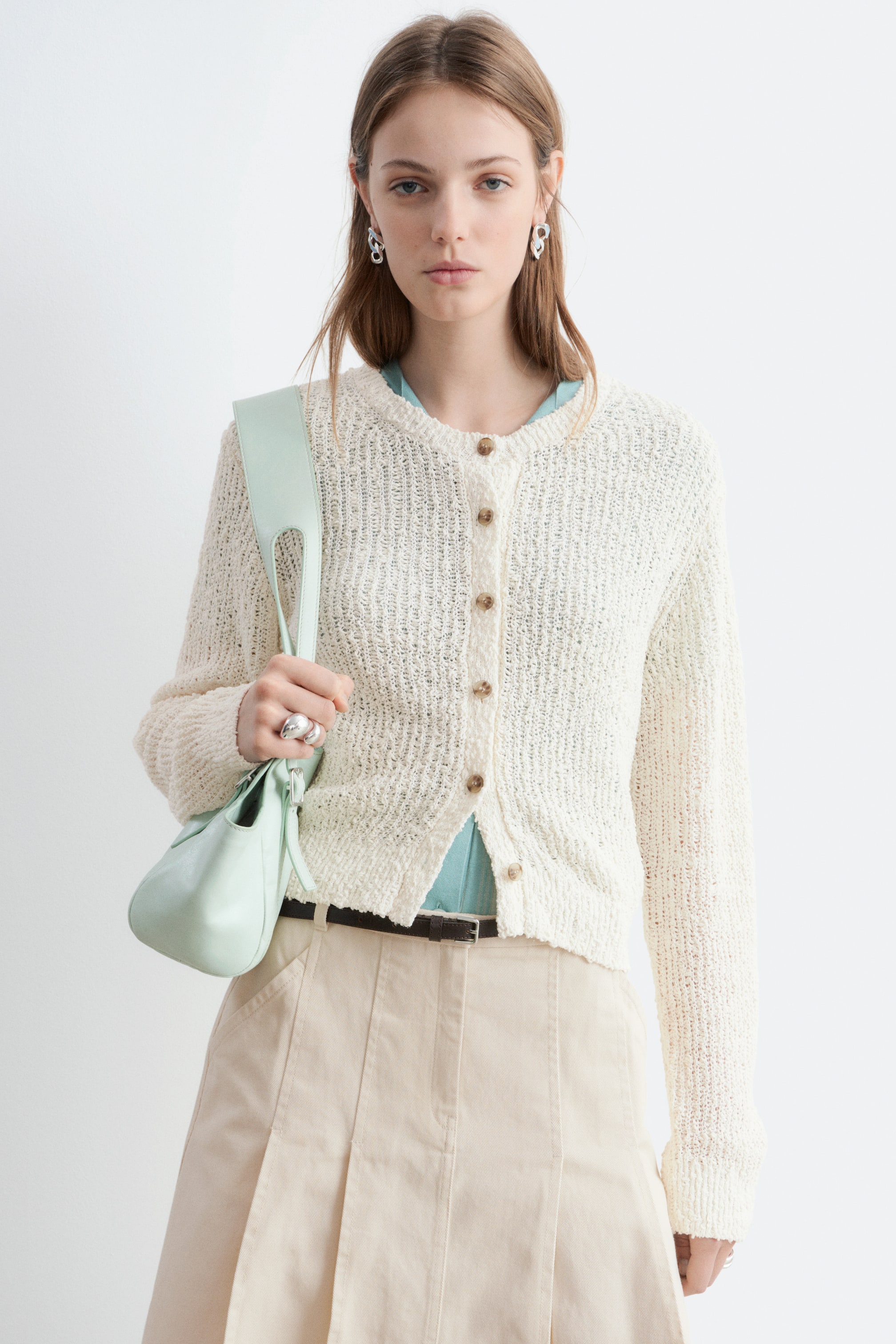 Zobraziť väčší obrázok: Nosí sa svetlo béžový otvorený pletený kardigan s okrúhlym výstrihom a dlhými rukávmi. Zapína sa vpredu hnedými gombíkmi a je vrstvený na svetlomodrom tope.