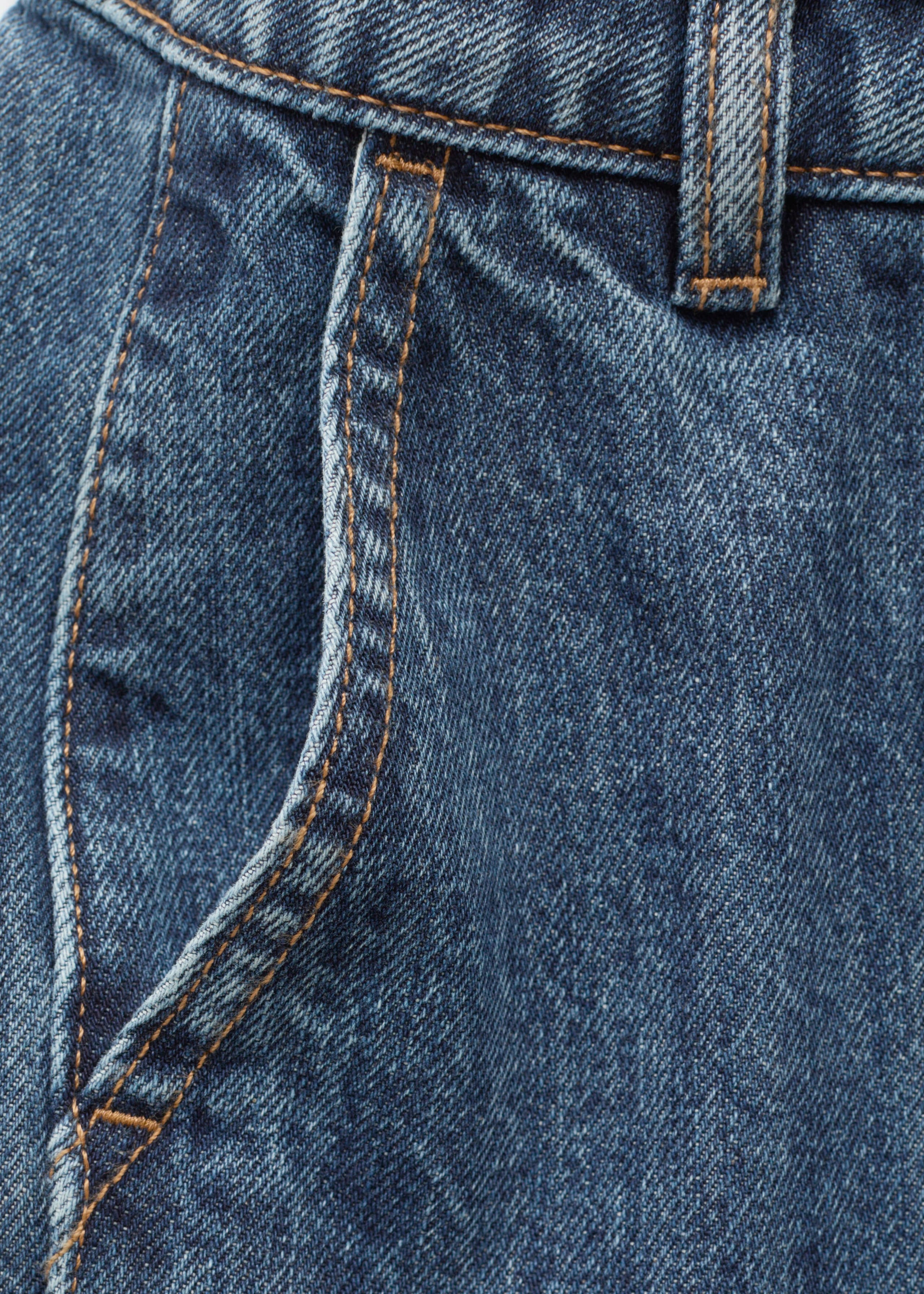 These blue denim jeans feature a close-up front view that highlights a slanted side pocket, a belt loop, and orange contrast stitching along the seams.