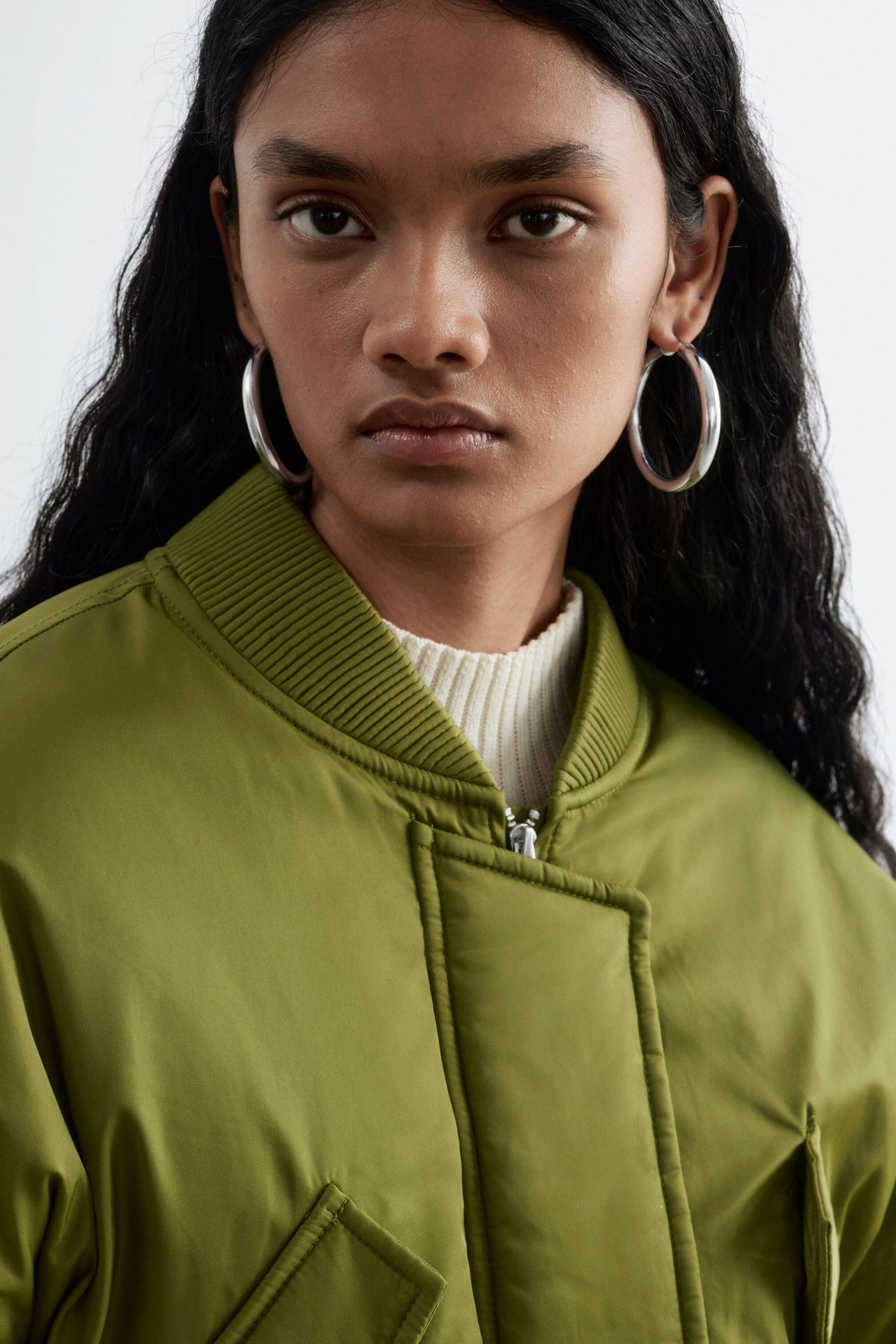 Ver imagen más grande: La modelo lleva unos pendientes de aro de tono plateado con acabado pulido. Los pendientes se combinan con una cazadora bomber verde y un cuello de punto canalé blanco.