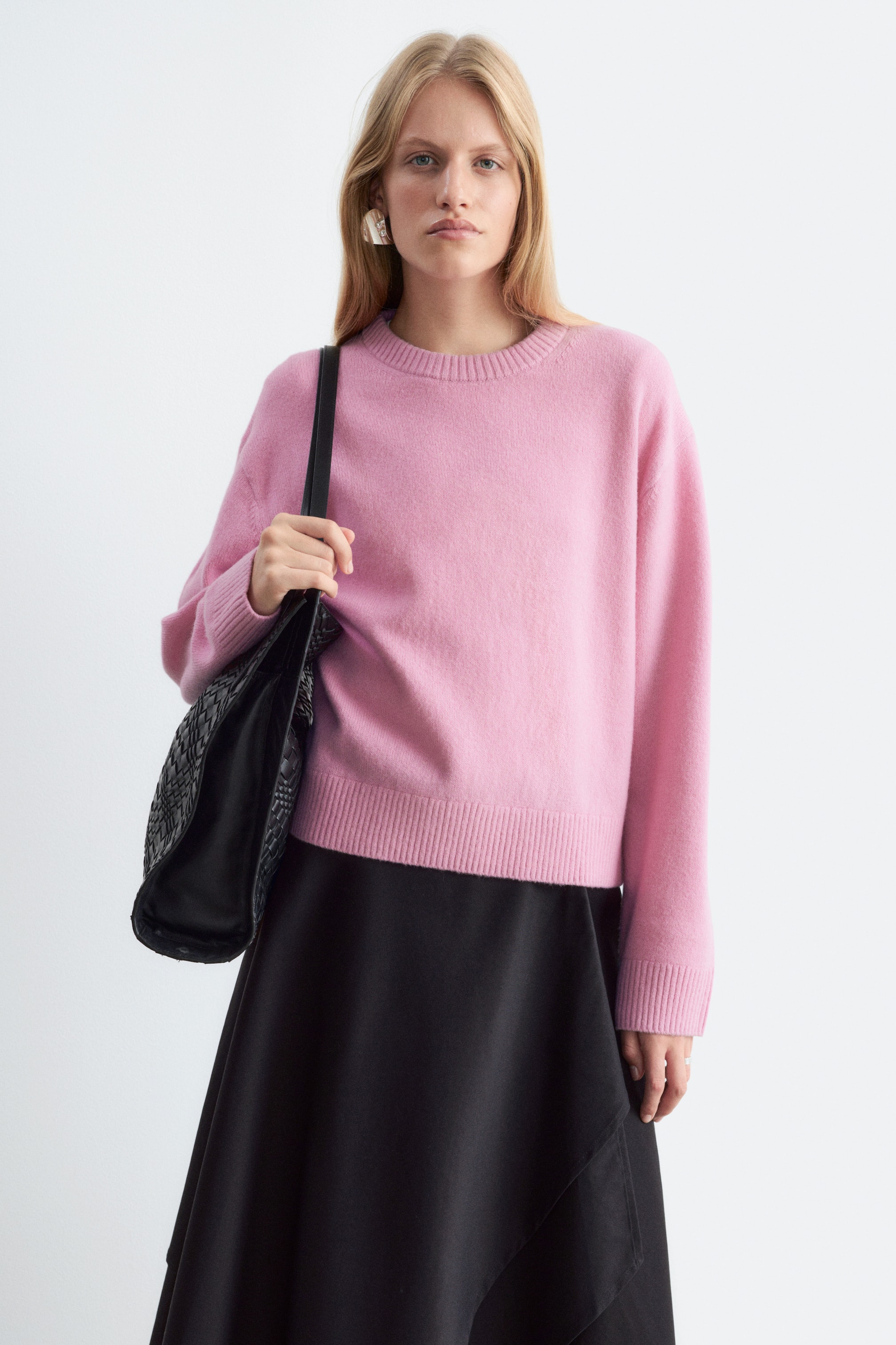 Agrandir l'image: Un pull en maille rose à coupe boxy, doté d’un col rond, de manches longues et de finitions côtelées, est porté par le mannequin. Le pull est associé à une jupe noire, et le mannequin porte un sac bandoulière tressé noir.