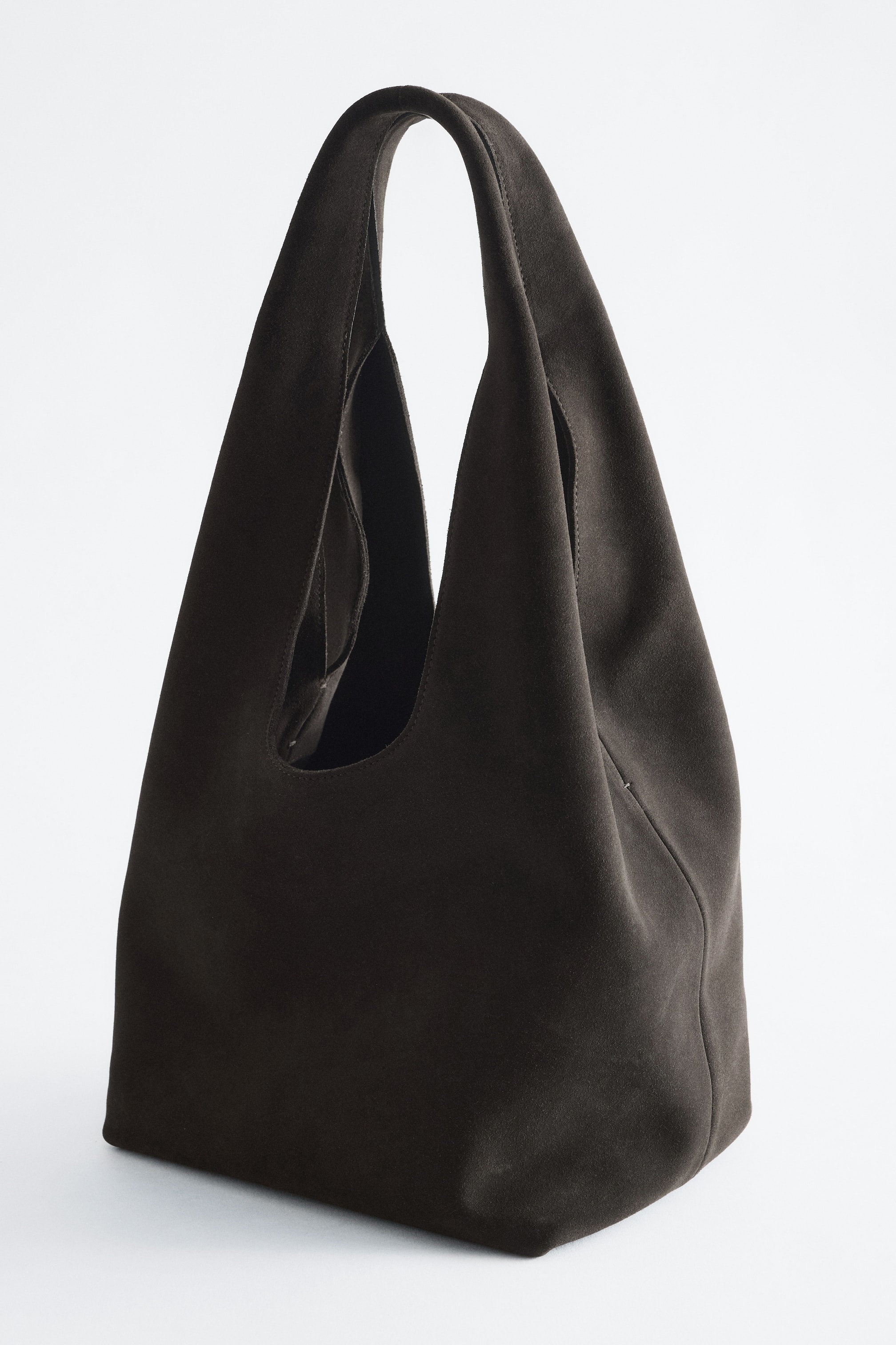 Vis større bilde: En mørkebrun totebag i delt semsket skinn står i en trekvart vinkel og viser frem sin romslige, slouchy silhuett og brede, integrerte skulderstropp. Vesken har en myk, matt tekstur med flat, strukturert bunn.