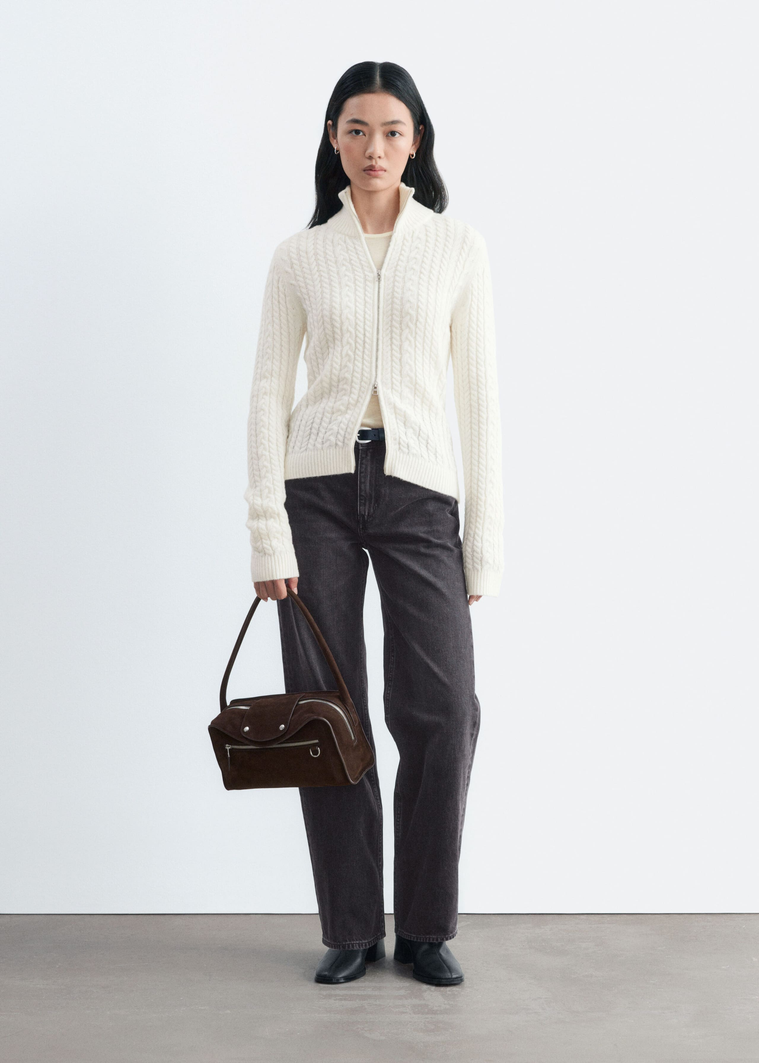 The model is wearing a cream cable-knit cardigan with a double-layer ribbed collar and a front zip. The cardigan is paired with dark grey jeans, and the model is holding a brown suede handbag.