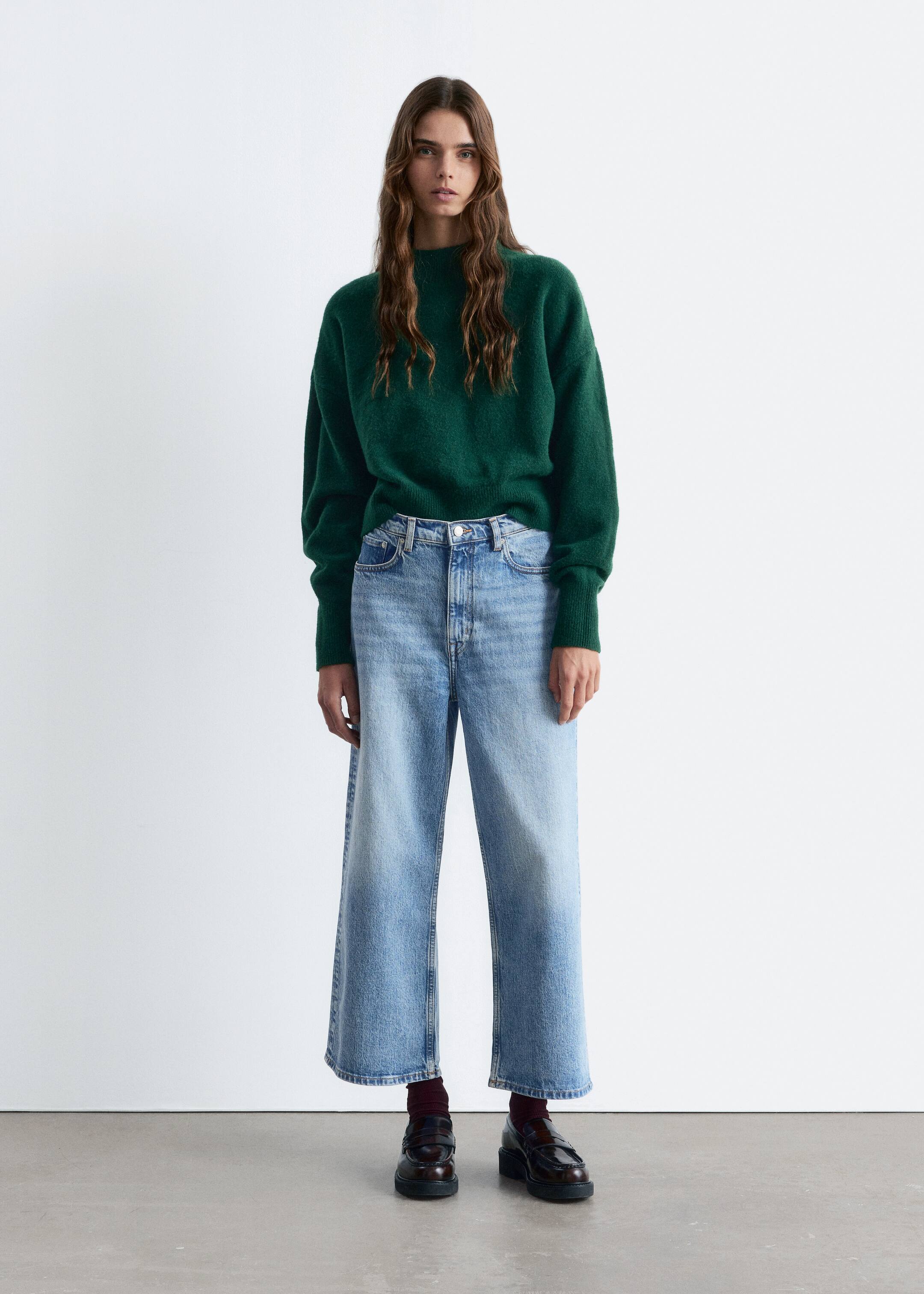 Visa större bild: En mörkgrön, avslappnad tröja med en hög krage och ribbade muddar bärs. Den är stylad med ljusa, högmidjade, vida jeans som slutar vid ankeln, och mörkbruna chunky loafers som avslöjar vinröda strumpor.