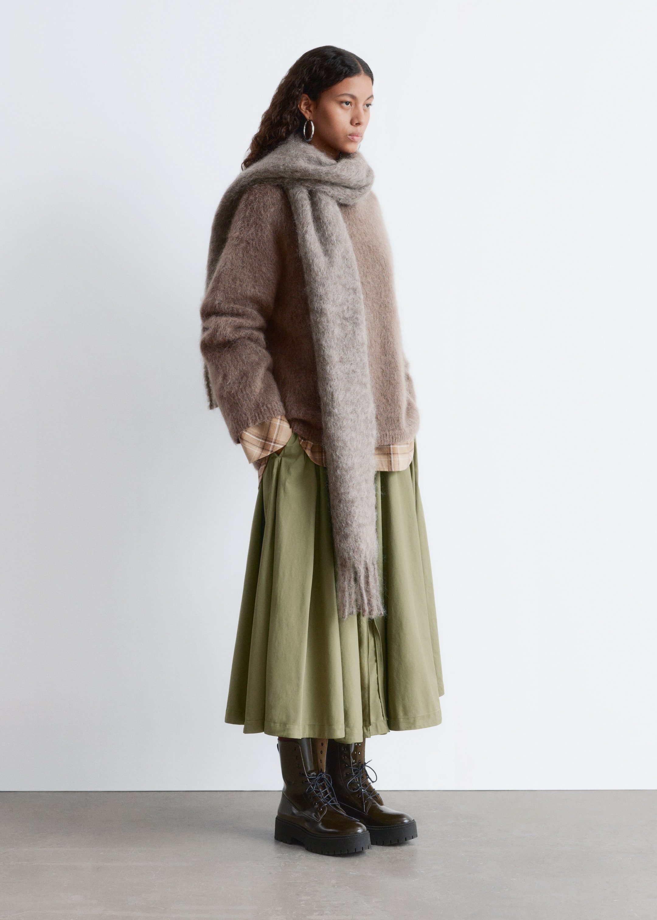 Ver imagem maior: Um traje com várias camadas está a ser exibido, mostrado de uma perspetiva angulada da esquerda. Apresenta um casaco castanho oversized e felpudo usado sobre uma camisa xadrez bege, uma saia midi verde-oliva, um longo cachecol cinza felpudo em volta do pescoço e botas de combate escuras com atacadores e solas grossas.