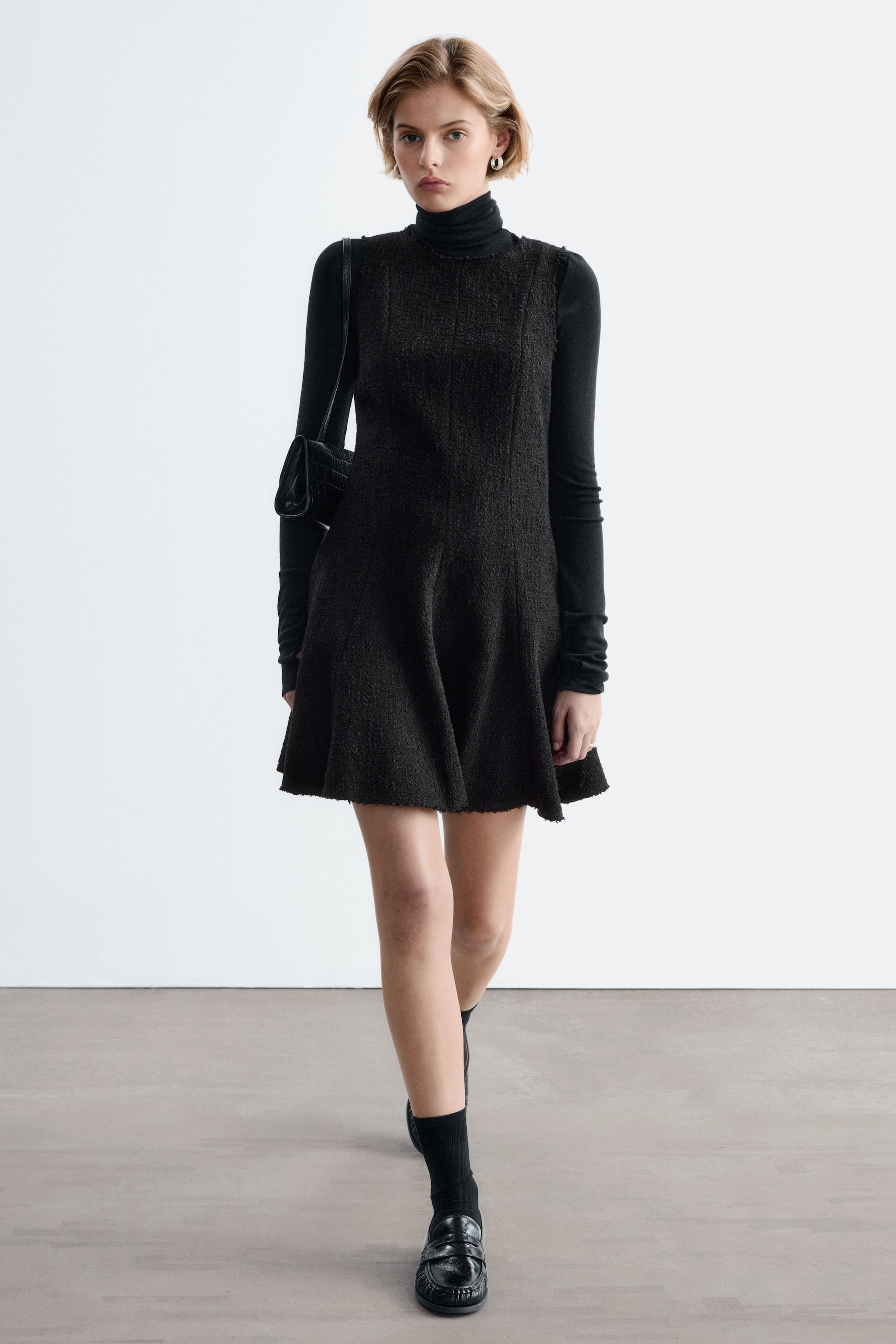 Agrandir l'image: Une mini-robe noire texturée à bords bruts et à silhouette évasée trapèze est portée par le mannequin. La robe sans manches est superposée à un top noir à col roulé et manches longues, et accessoirisée avec un petit sac bandoulière noir.