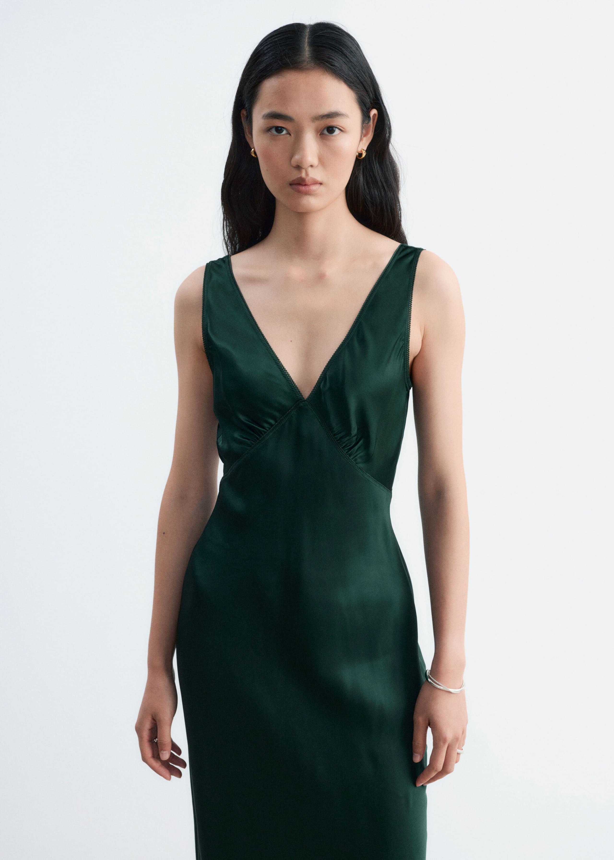 A dark green satin slip dress featuring a V-neckline and a softly gathered bust is worn by the model. The sleeveless dress drapes into a clean, fitted silhouette.
