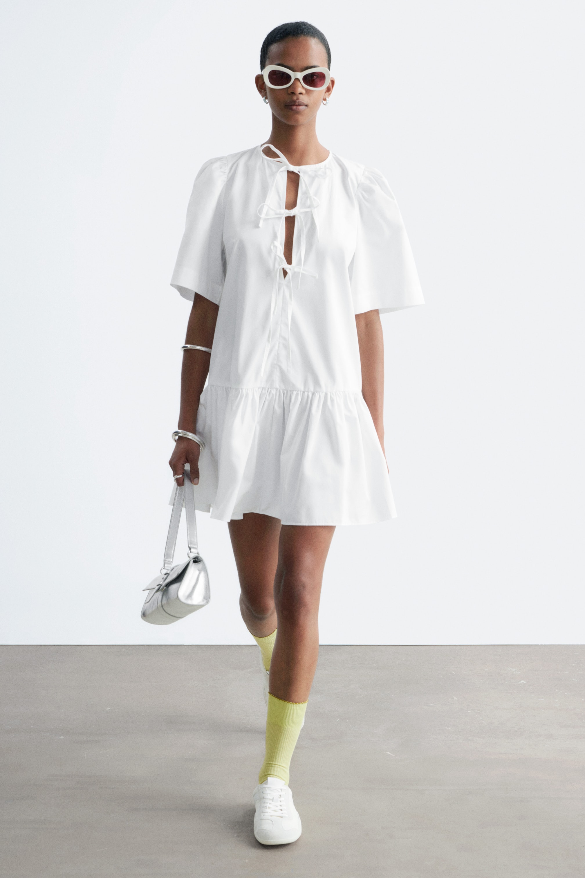 Grotere afbeelding bekijken: Een witte mini-jurk met een ronde halslijn en zelfbindende sluitingen aan de voorkant, korte pofmouwen en een geplooide verlaagde taille die leidt naar een uitlopende rok.