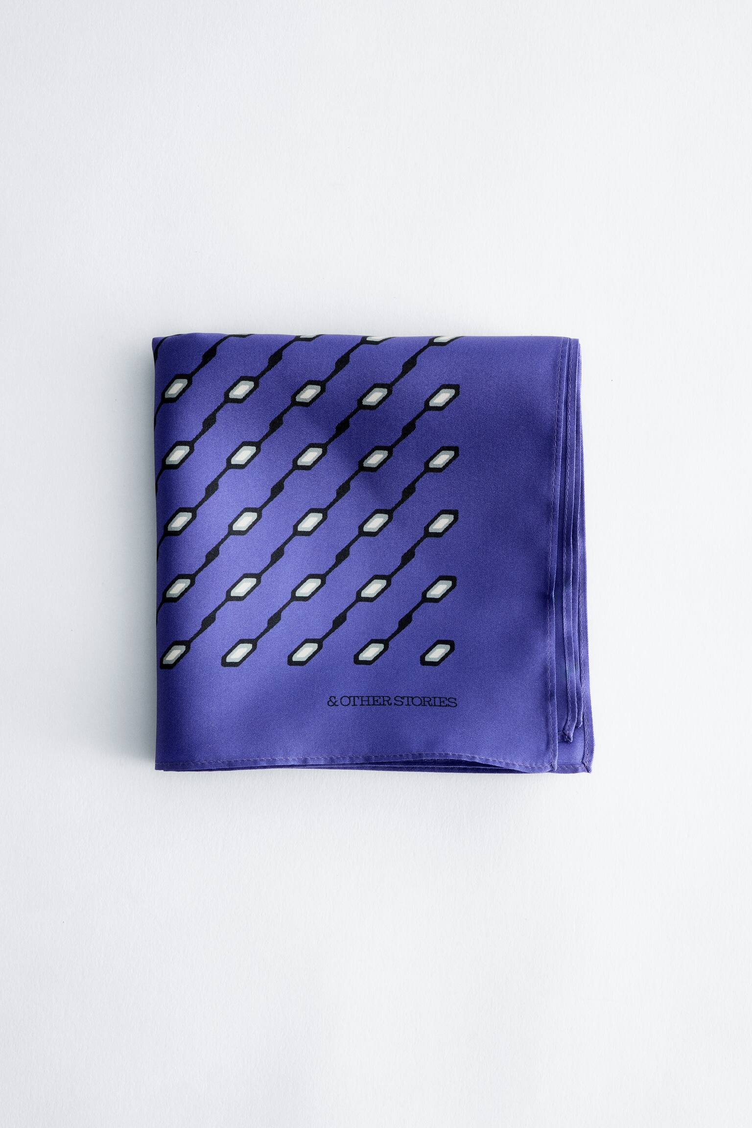 Een paarse zijden sjaal met print ligt plat en opgevouwen, met de voorkant naar voren, en toont een diagonaal geometrisch patroon van zwarte lijnen met kleine witte en grijze zeshoekige vormen. De merknaam "& OTHER STORIES" staat in zwart gedrukt bij de rechteronderhoek, en de sjaal is afgewerkt met netjes gestikte, gerolde randen.