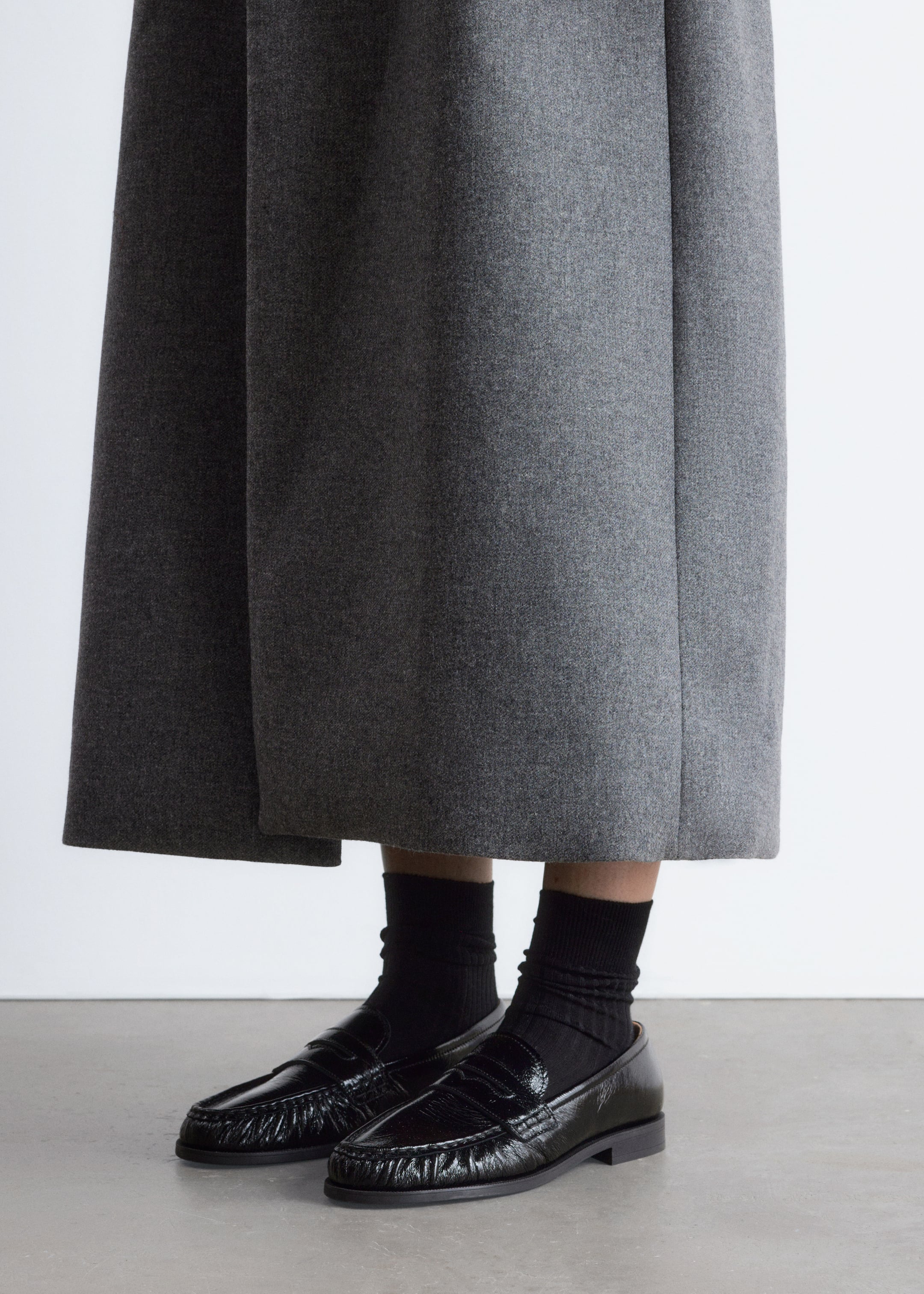 Ver imagen más grande: Un par de mocasines de charol negro está siendo exhibido desde un ángulo lateral ligeramente elevado, con costuras estilo mocasín en la puntera y un tacón bajo de bloque. Los mocasines se llevan con calcetines negros de punto y pantalones grises de pierna ancha, que caen justo por encima del tobillo.