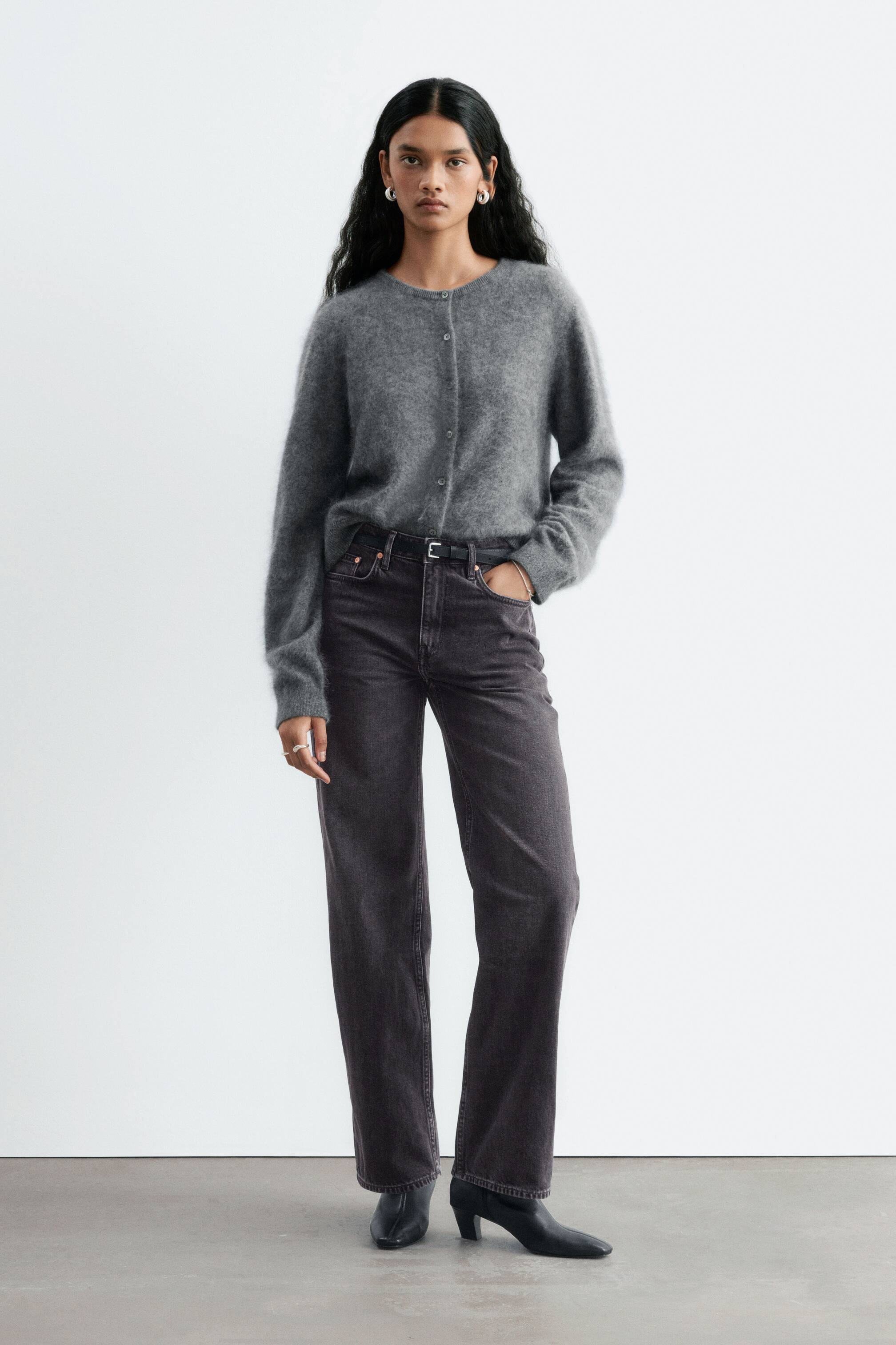 Agrandir l'image: Le mannequin porte un jean droit gris foncé à taille mi-haute, en position debout avec une main glissée dans la poche avant. Le jean est associé à une fine ceinture noire, un cardigan en maille gris et des bottes noires à talons.