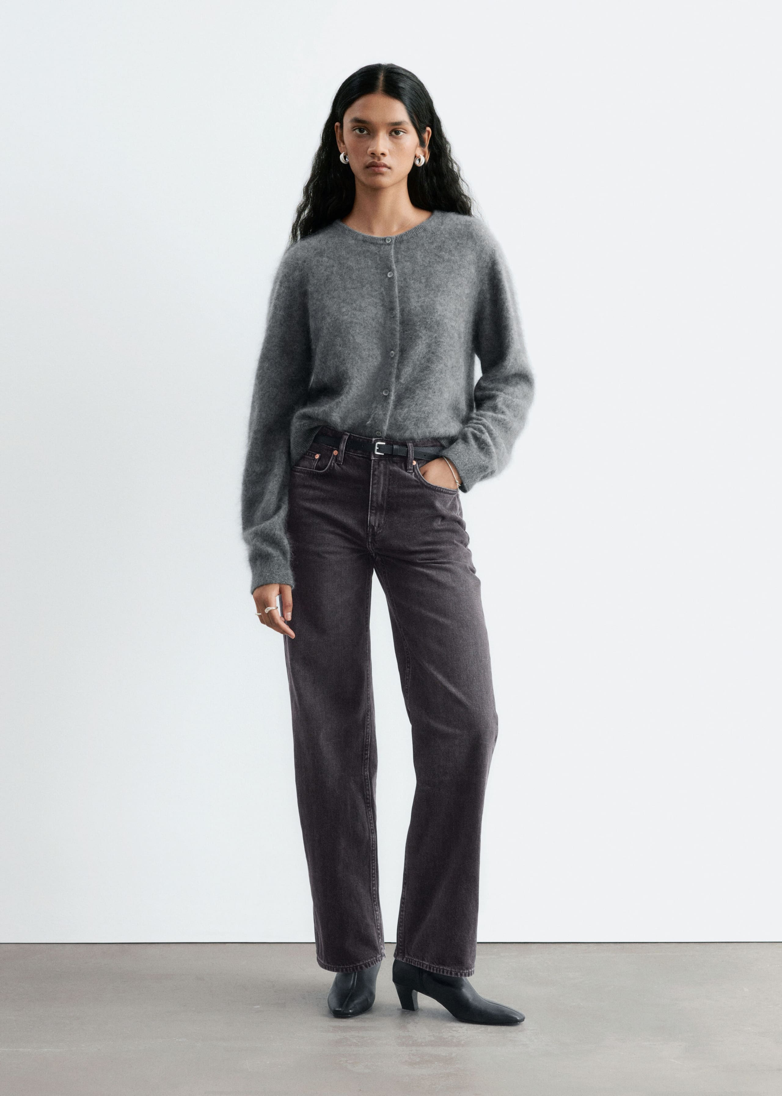 Dark gray straight-leg jeans with a mid-rise waist are being worn by the model, who is standing with one hand resting in a front pocket. The jeans are styled with a thin black belt, a gray knit cardigan, and black heeled boots.