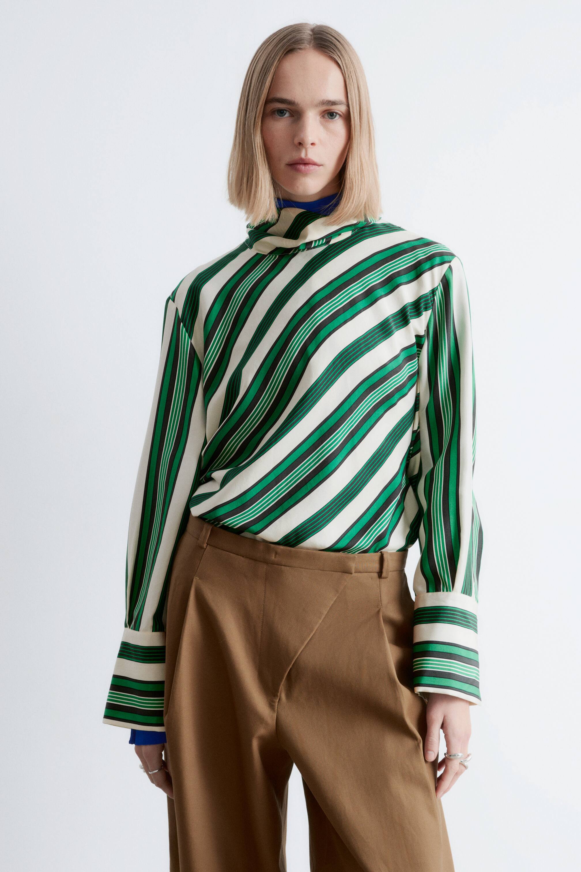 Agrandir l'image: Une blouse rayée est orientée vers l'avant, avec un fond crème orné de rayures diagonales vertes et fines noires. Elle est conçue avec un col drapé haut, dévoilant un sous-vêtement bleu, et possède des manches longues se terminant par de larges poignets rayés, semblant rentrées dans un pantalon marron.