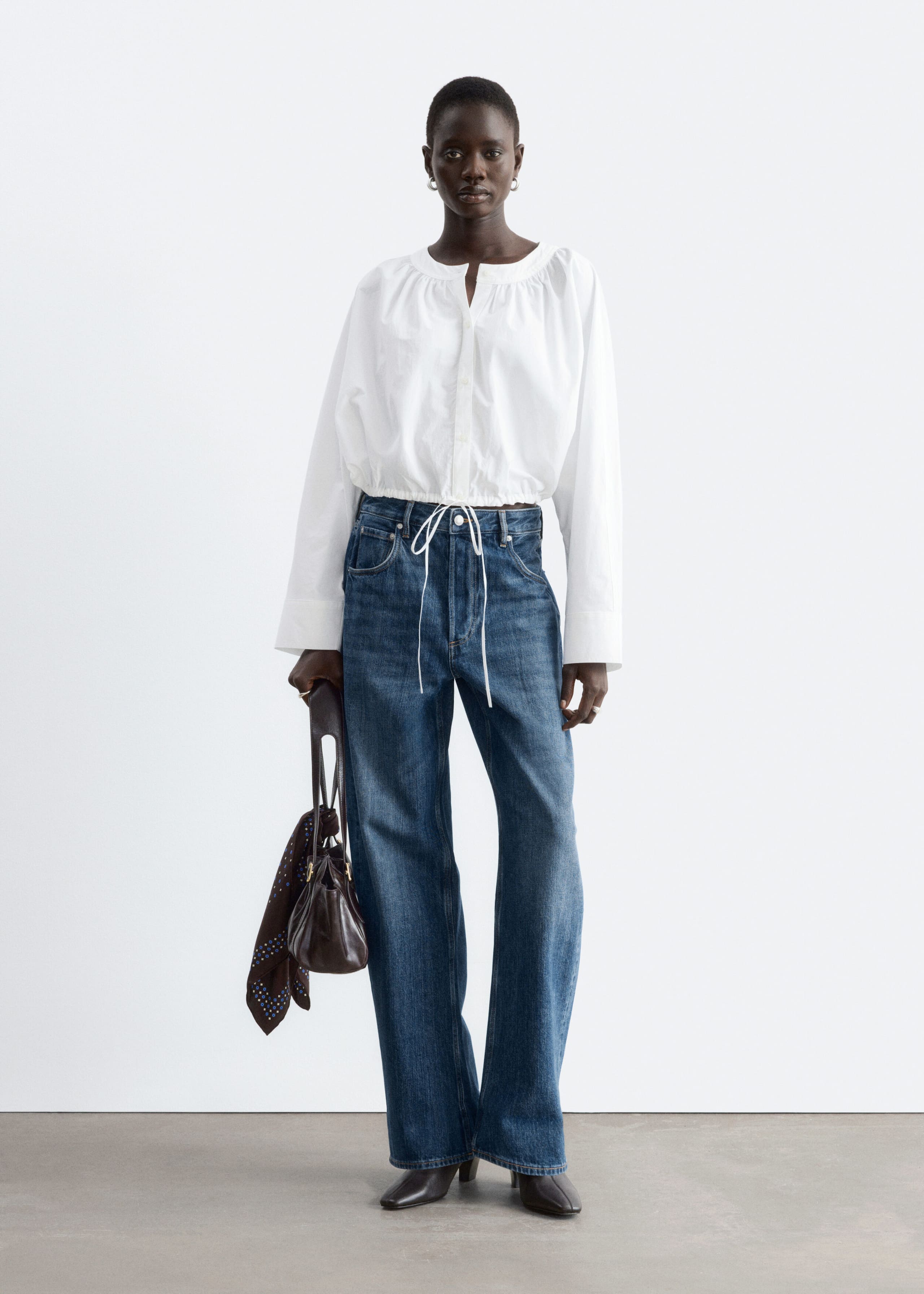A white cropped long-sleeve blouse featuring gathered detailing around the neckline, a buttoned placket, and a tied drawstring hem is worn by the model. The blouse is styled with wide-leg blue jeans, and the model is carrying a dark brown handbag.