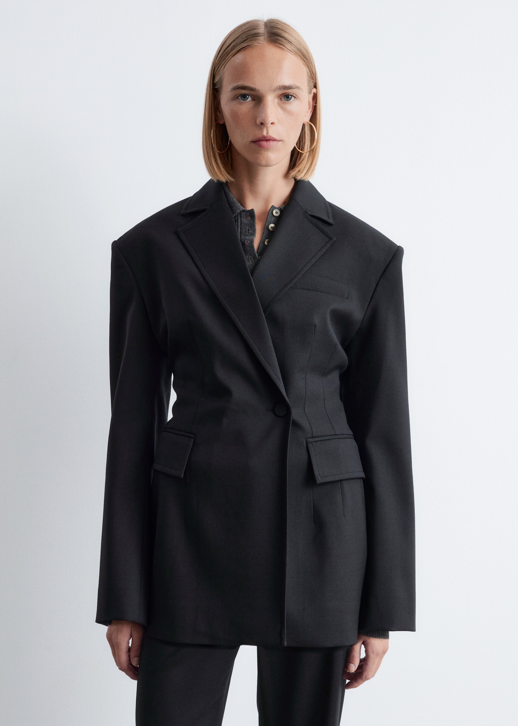 Agrandir l'image: Un blazer noir ajusté est porté, légèrement incliné vers la droite, avec de larges revers crantés, une fermeture à un bouton et des épaules structurées proéminentes. Le blazer est cintré à la taille avec des coutures verticales et comprend deux poches à rabat, superposé à un t-shirt Henley gris.