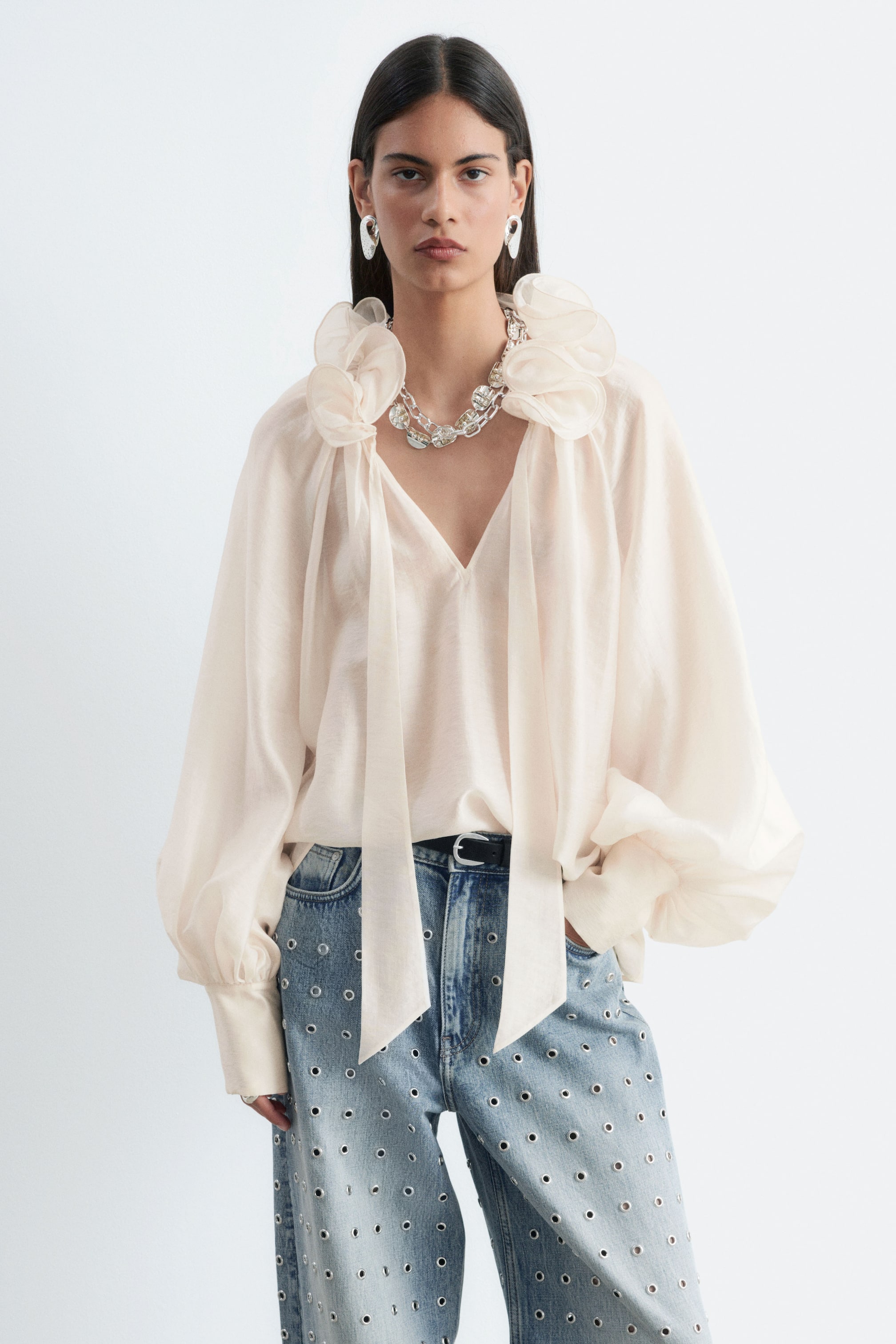 Grotere afbeelding bekijken: Een crèmekleurige, semi-transparante, volumineuze blouse met een gerimpelde kraag met lange striklinten, een V-hals en wijd vallende ballonnenmouwen wordt gedragen door het model. De blouse is gestyled ingestopt in lichtgewassen blauwe jeans, versierd met zilverkleurige ringen.
