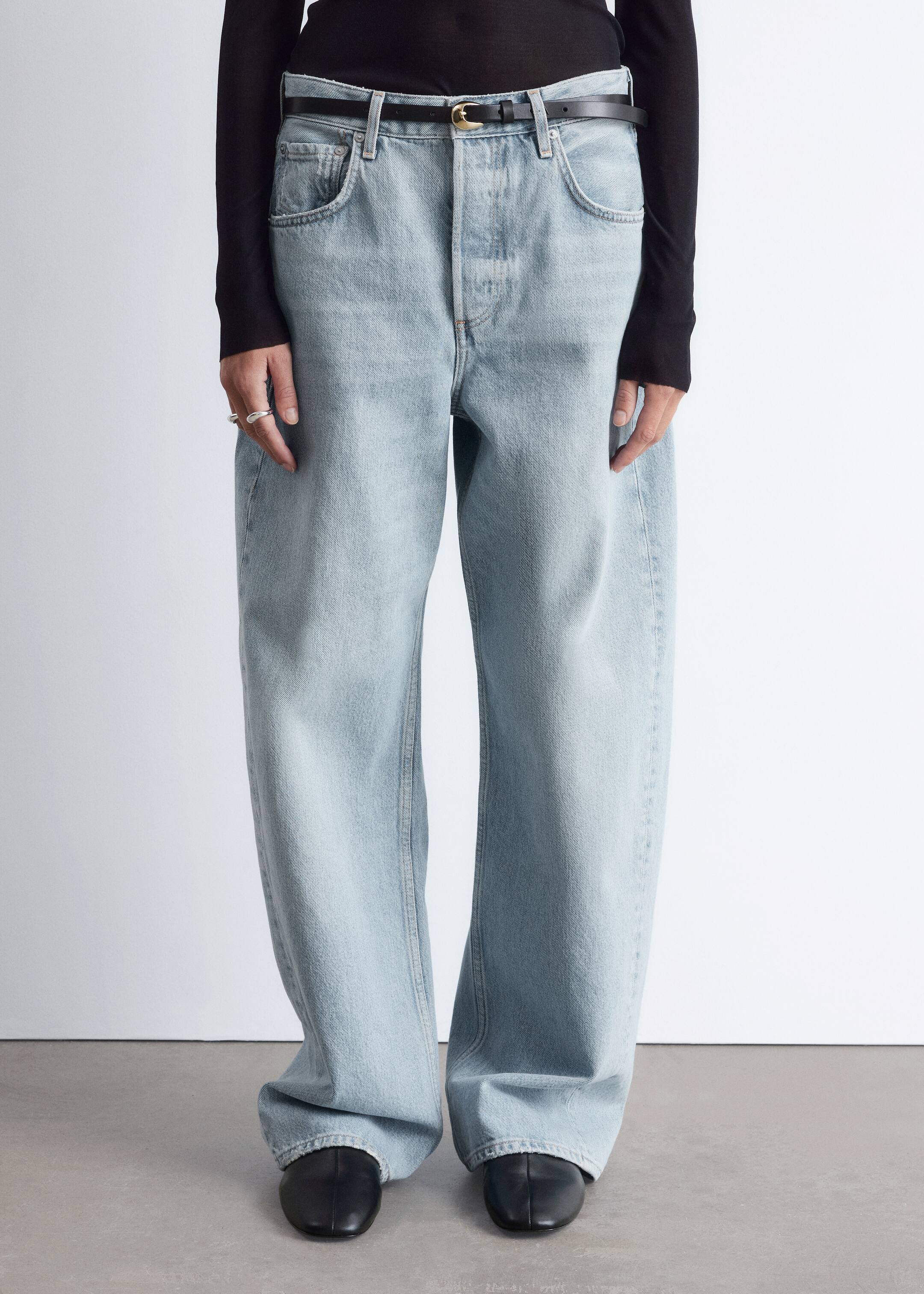 View larger image: Light blue, wide-leg jeans are being presented, featuring a relaxed fit, a visible button fly, and front scoop pockets. A thin black belt with a gold buckle is cinching the waist, and the full-length denim is pooling over black shoes.