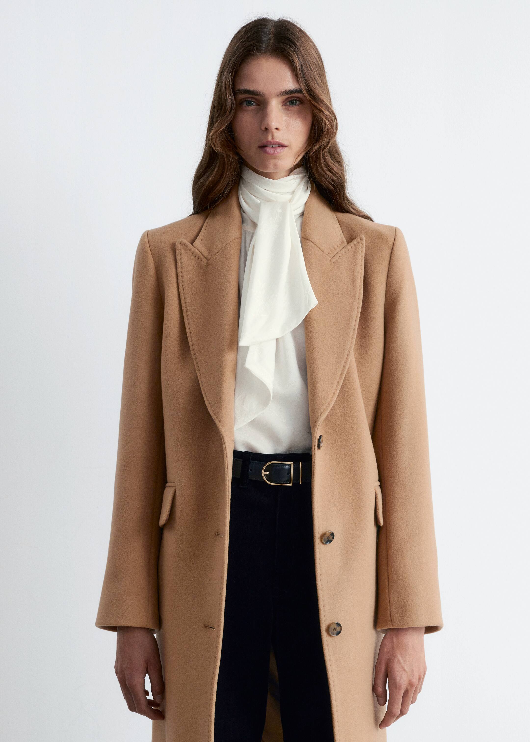 View larger image: A camel coat is being worn, featuring visible stitching on its wide notch lapels, a single-breasted front with two brown buttons, and flap pockets. It is styled over a cream pussy-bow blouse and black trousers.
