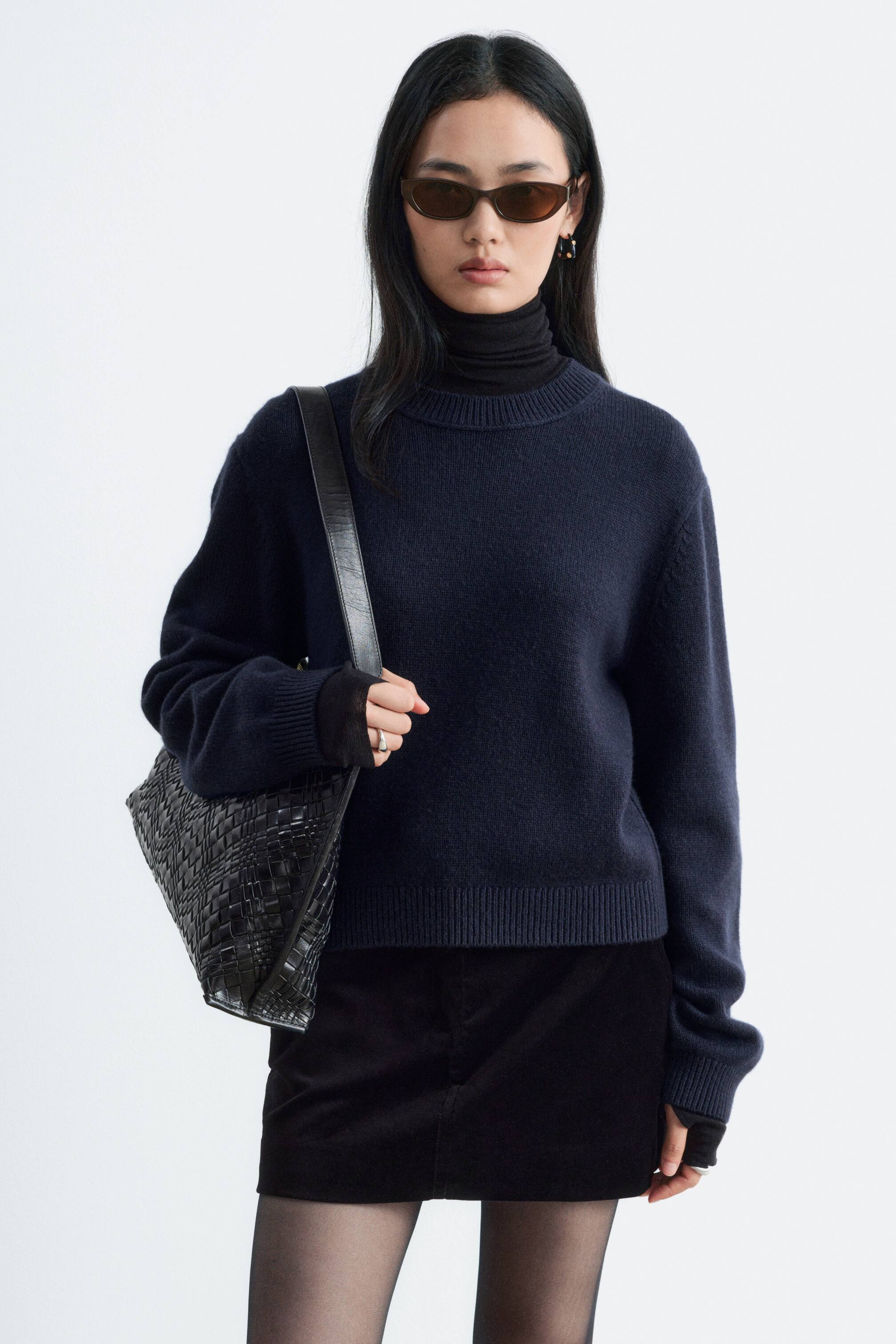 Größeres Bild anzeigen: Ein dunkelmarineblauer Pullover aus Kaschmir-Mischung mit Rundhalsausschnitt und Rippbündchen wird vom Model über einem schwarzen Rollkragenpullover getragen. Der Pullover ist mit einem schwarzen Rock kombiniert, und das Model trägt eine schwarze Schultertasche aus geflochtenem Leder.